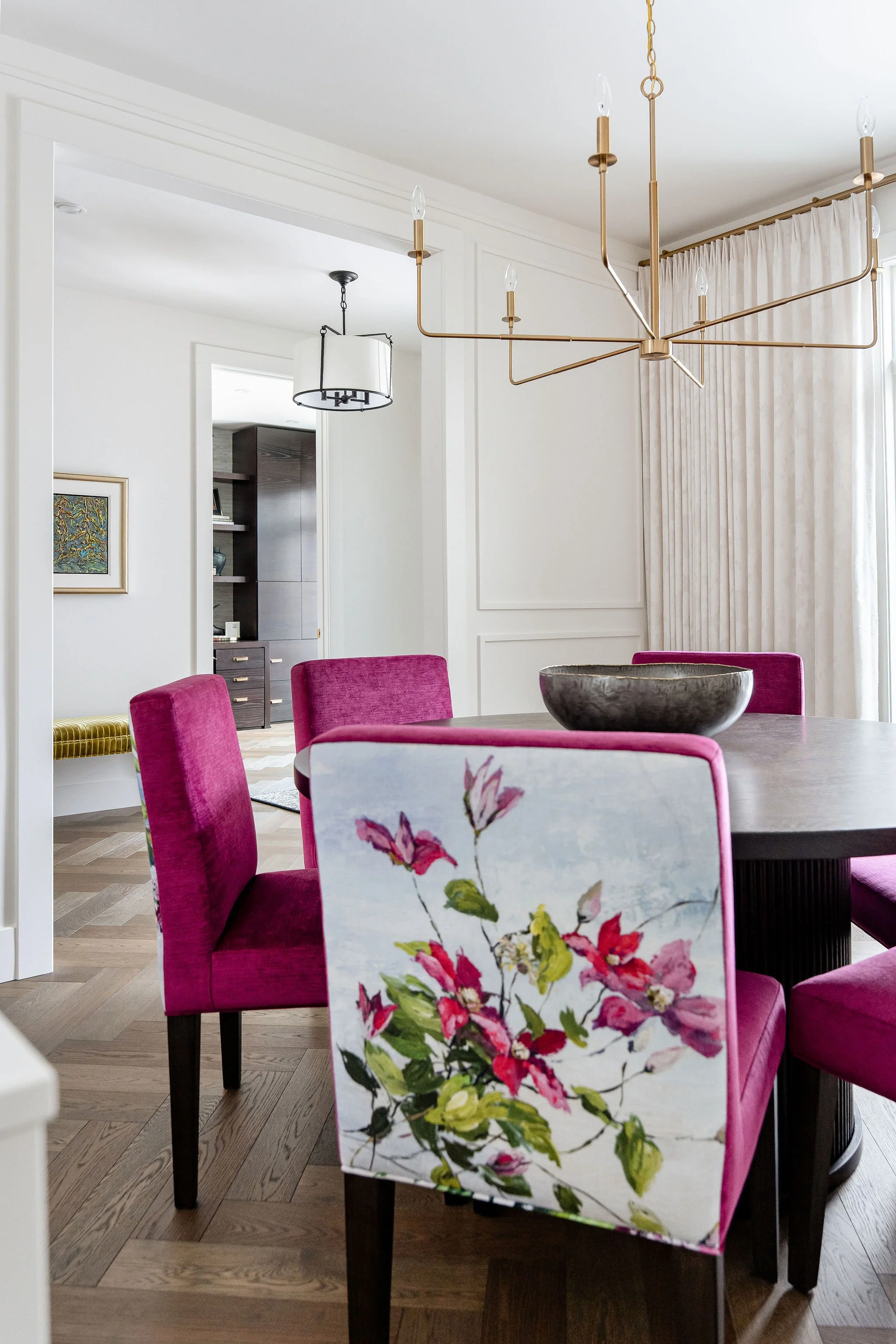 A dining room with a round table and pink velvet chairs. A floral-patterned chair is visible in the foreground. There is a modern chandelier hanging above the table. The room has white walls, light wood flooring, a window with cream-colored curtains,