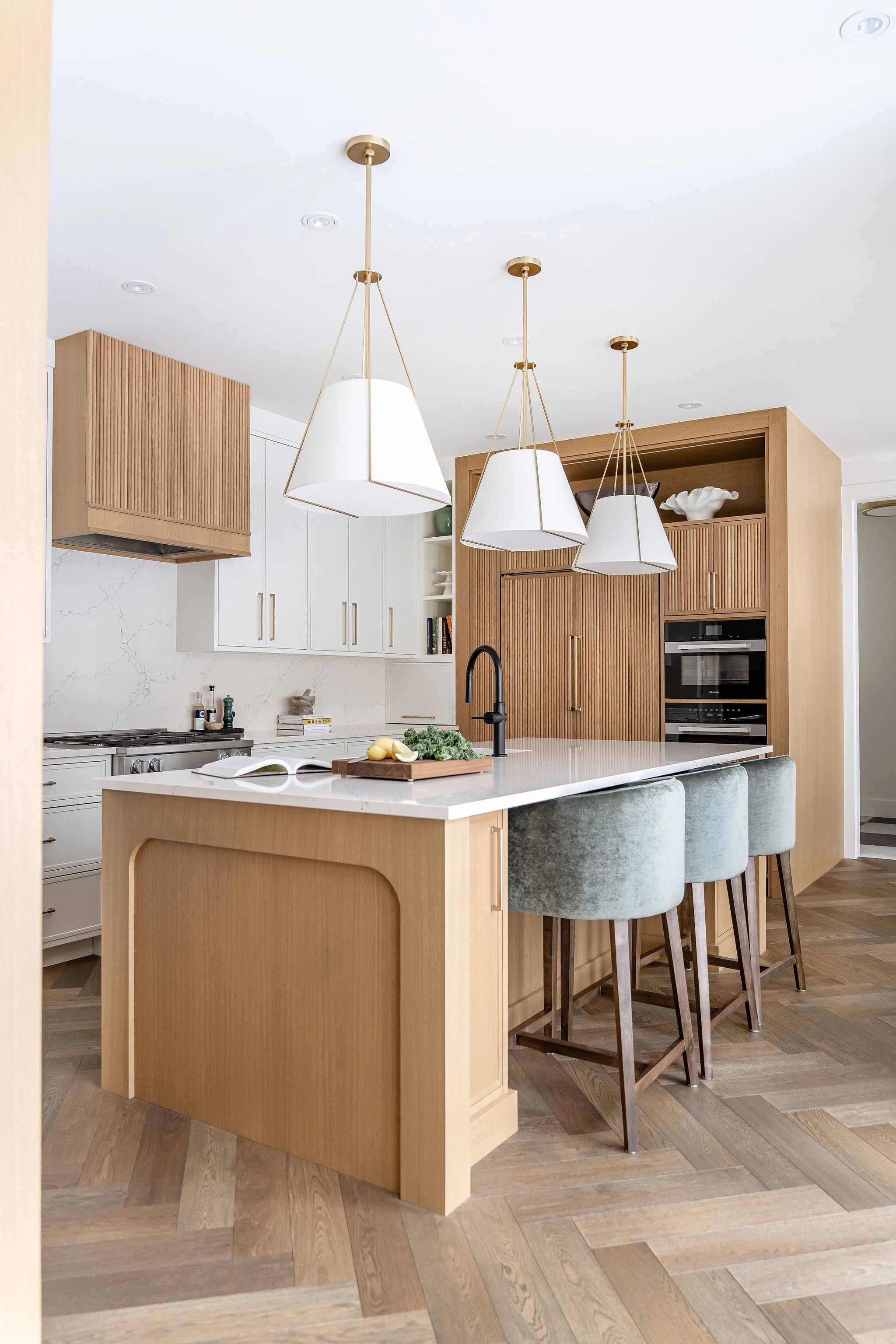 Modern kitchen with a white marble island, four green upholstered bar stools, wooden cabinets, white and natural wood finish, pendant lights, and a built-in oven.