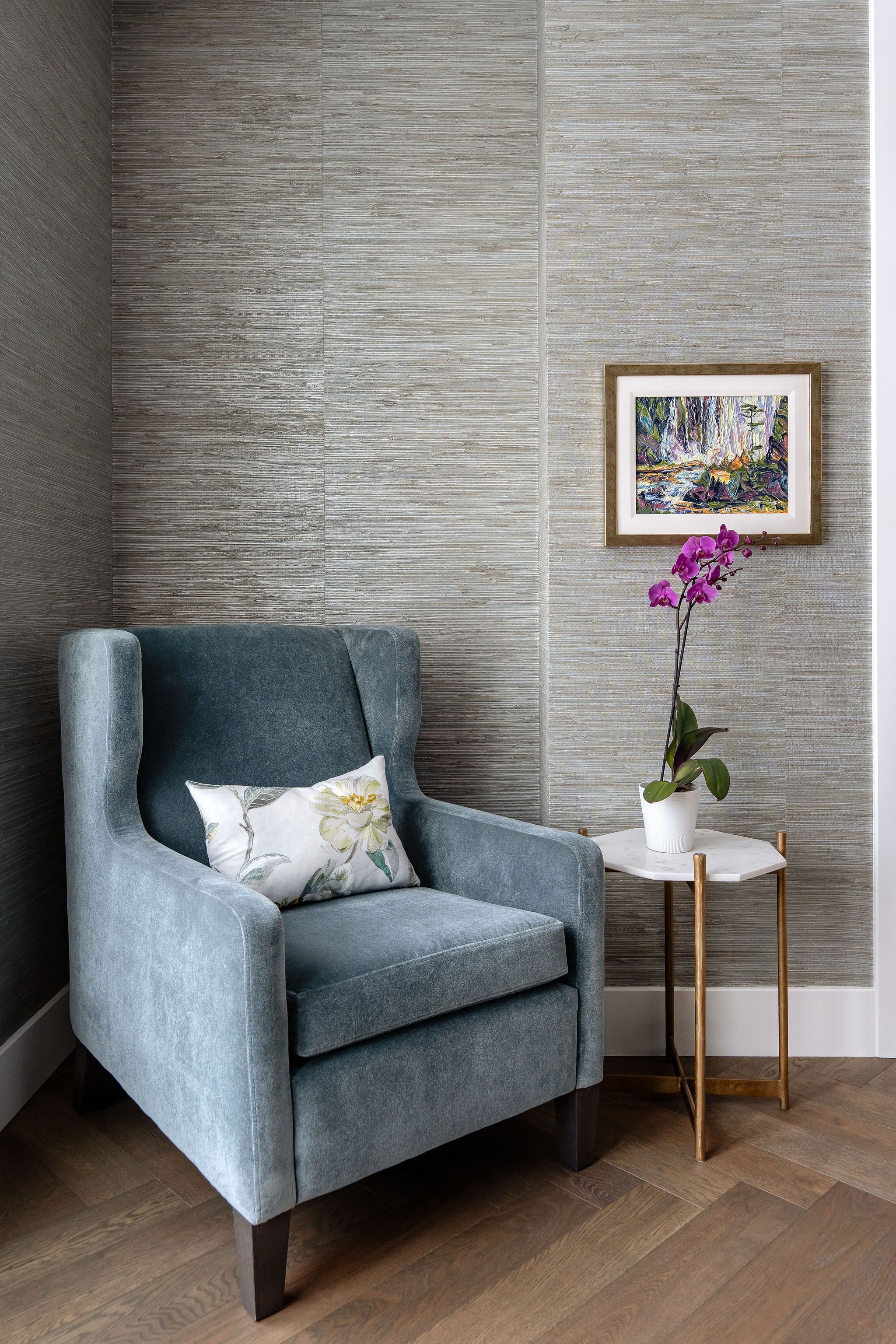 A cozy living room corner with a gray armchair, a small side table with a potted orchid, and framed artwork on textured beige walls.