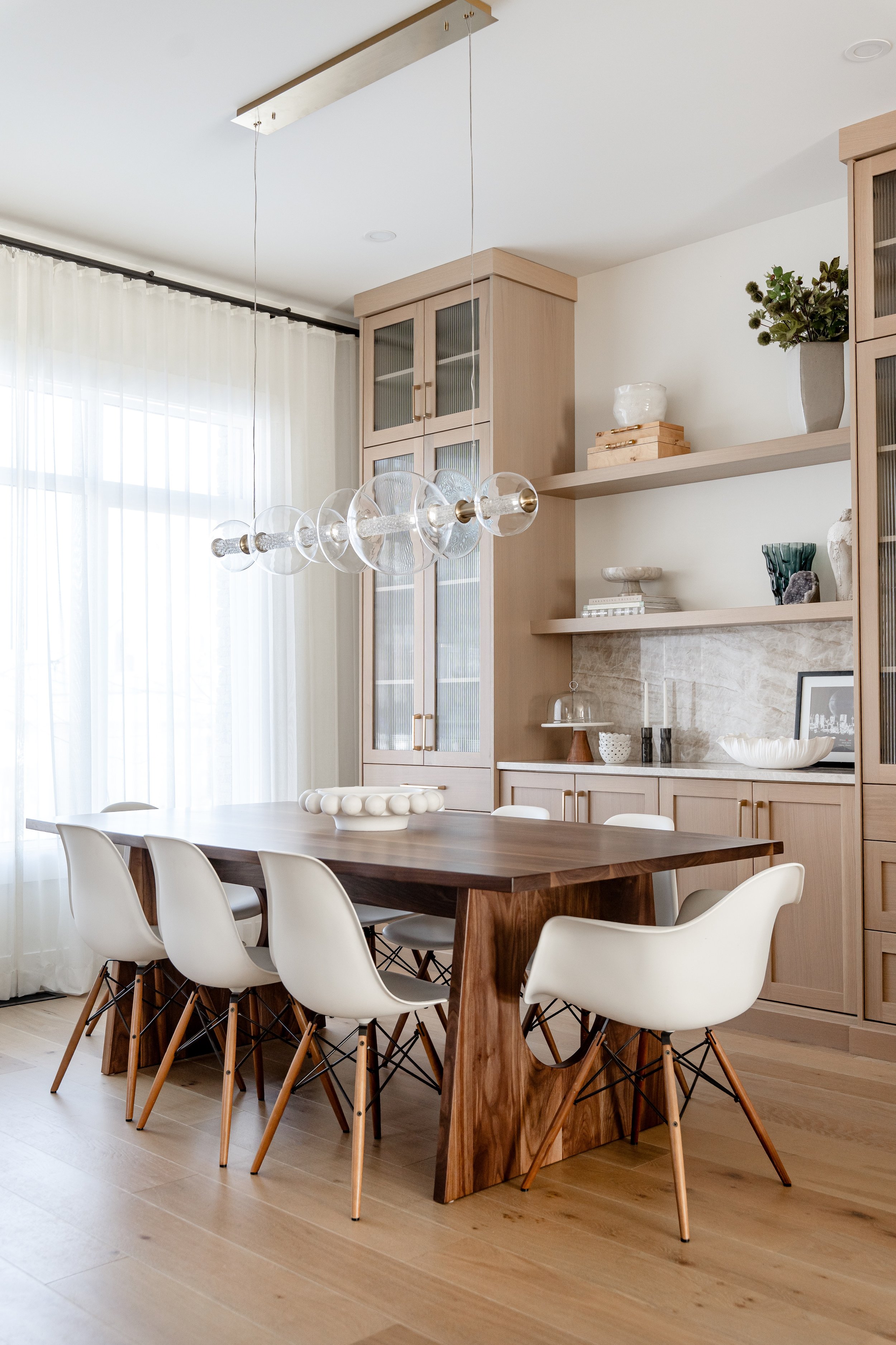 Modern dining room with a large wooden table, white chairs, a glass bubble chandelier, built-in wooden cabinets, open shelves with decorative items, and large windows with sheer curtains.