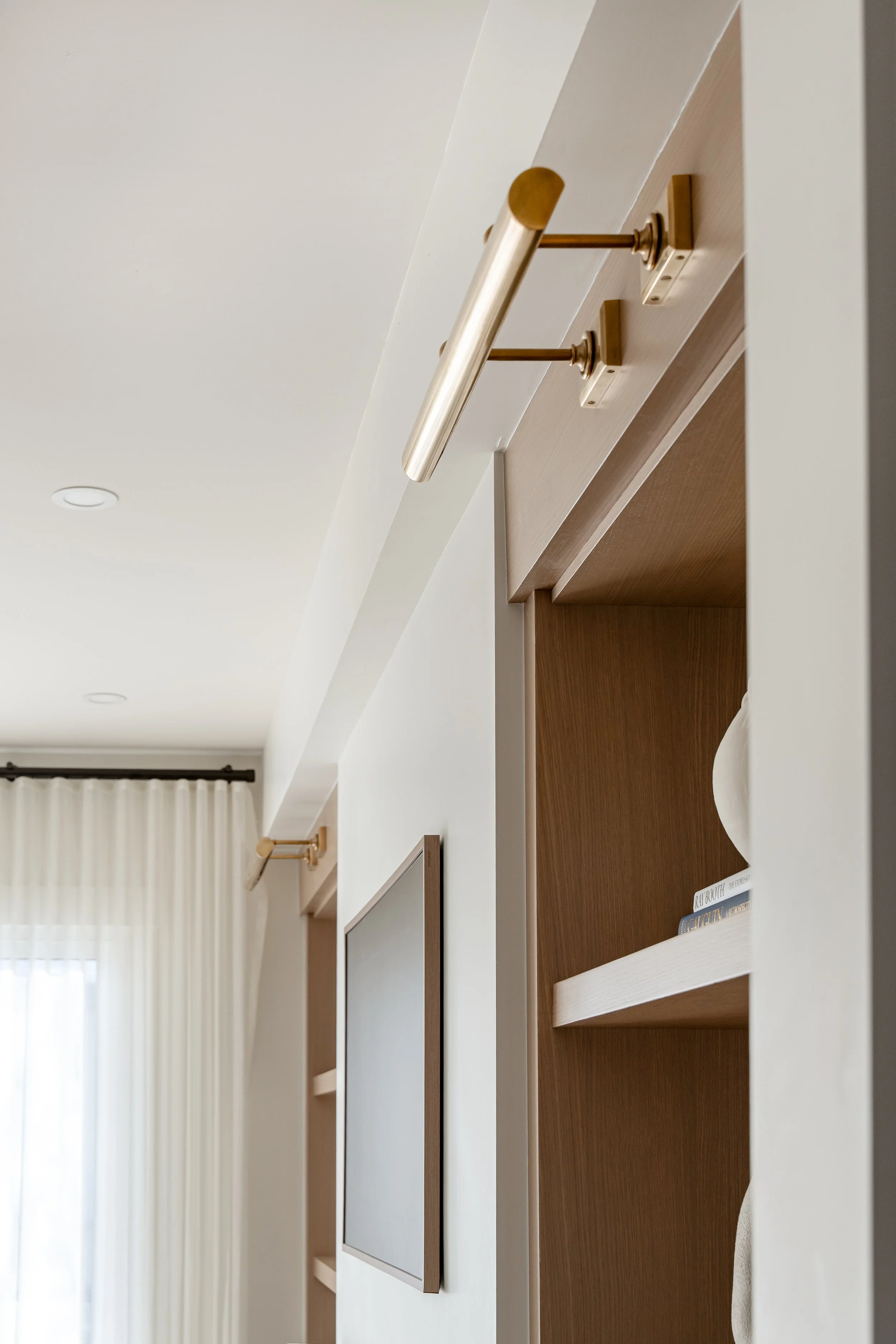 Interior view of a room with a modern wall-mounted light fixture, a flat-screen TV, wooden shelving, and a window with white curtains.