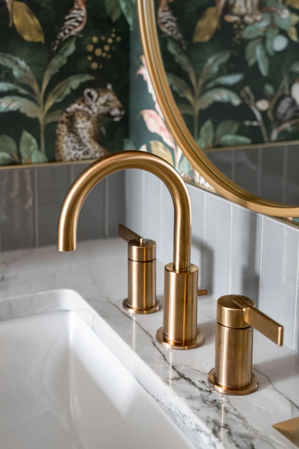 Close-up of a gold faucet and handles above a white sink, with a mirror reflecting a wall mural featuring a leopard and leafy plants.