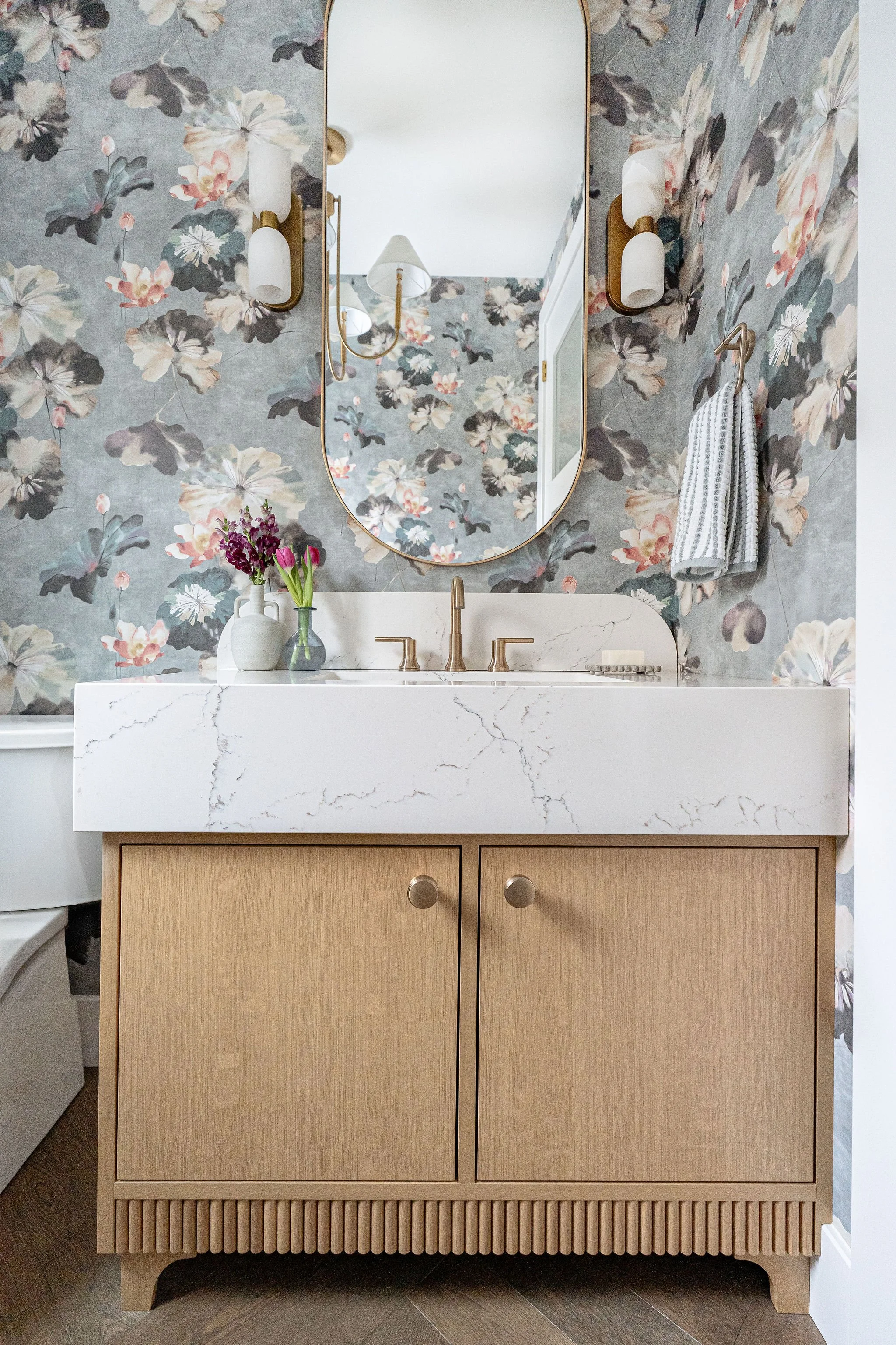 Bathroom vanity with a marble countertop, a large oval mirror, a vase of purple flowers, and wooden cabinetry with slatted bottom design.