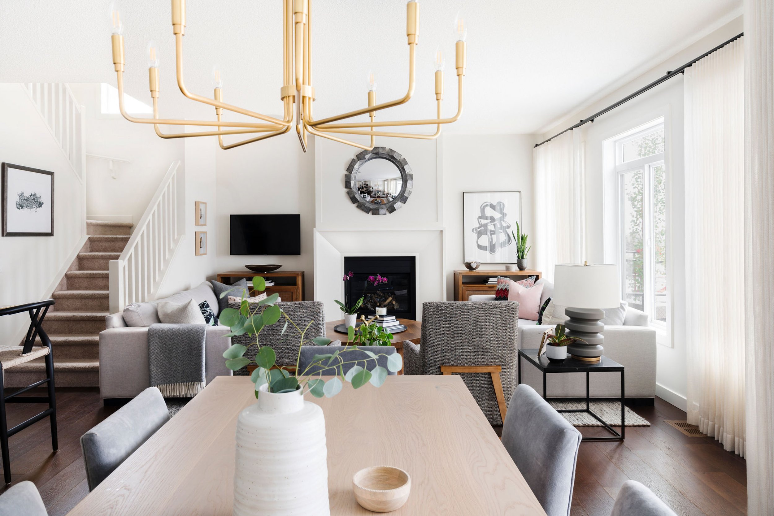 Modern living room with beige and gray furniture, wooden dining table, gold chandelier, fireplace, and large windows with white curtains. Decor includes potted plants, artwork, and a round mirror above the fireplace.