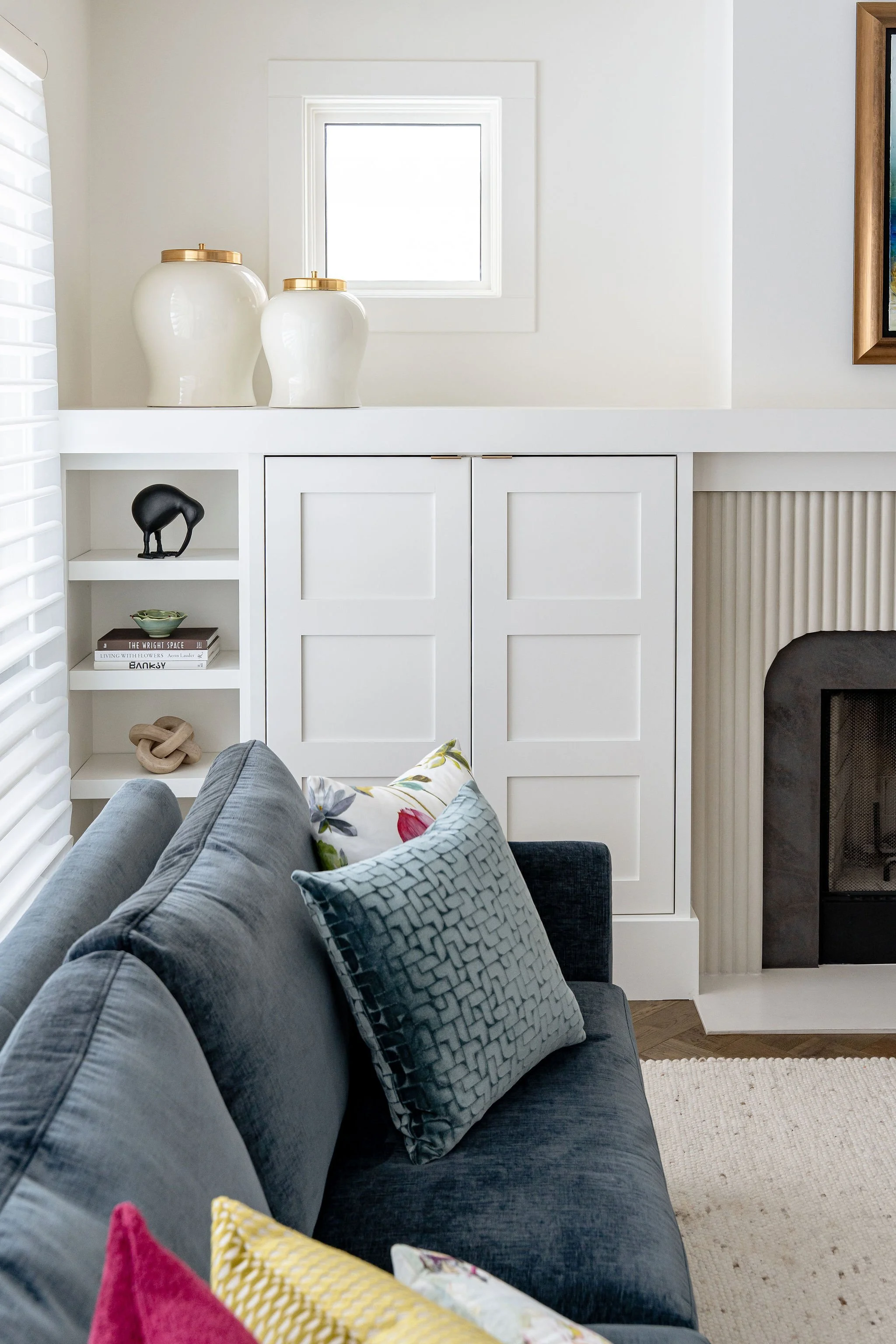 Living room with gray sofa, decorative pillows, white cabinetry, black sculpture, ceramic vases on shelf, small window, partially visible fireplace, wall art, and a woven rug.