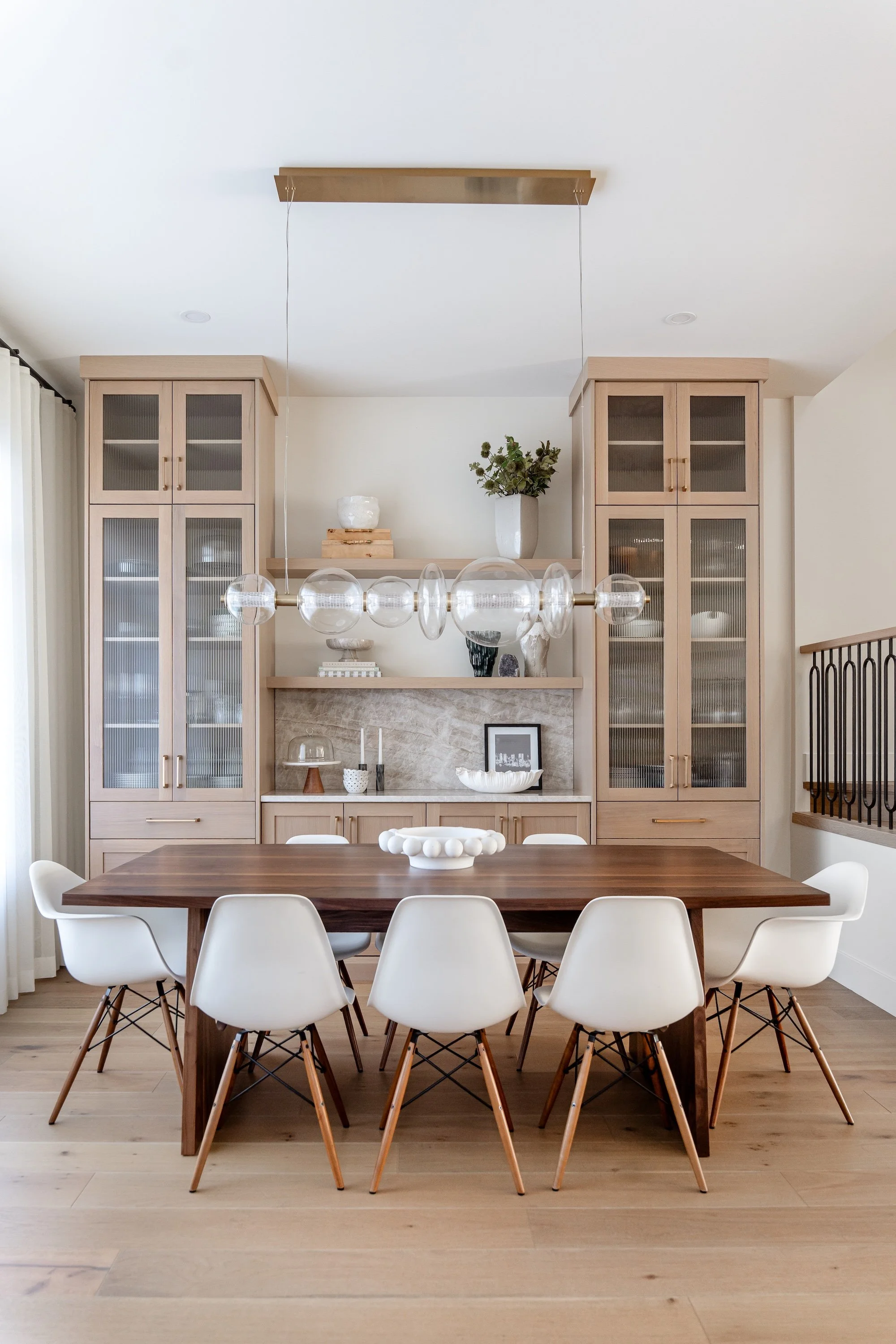 A dining room with a wooden table surrounded by white chairs with wooden legs, a built-in wooden cabinet with glass doors, a modern chandelier with large glass globe pendants, and decorative items such as vases, candles, and a framed photo on a marbl