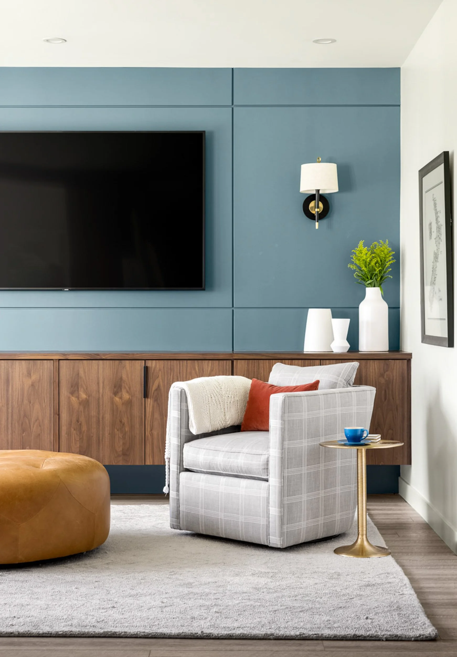 Modern living room with blue accent wall, wall-mounted TV, wooden cabinet, gray plaid armchair, leather ottoman, gold side table with blue cup, and decorative vases and plants.