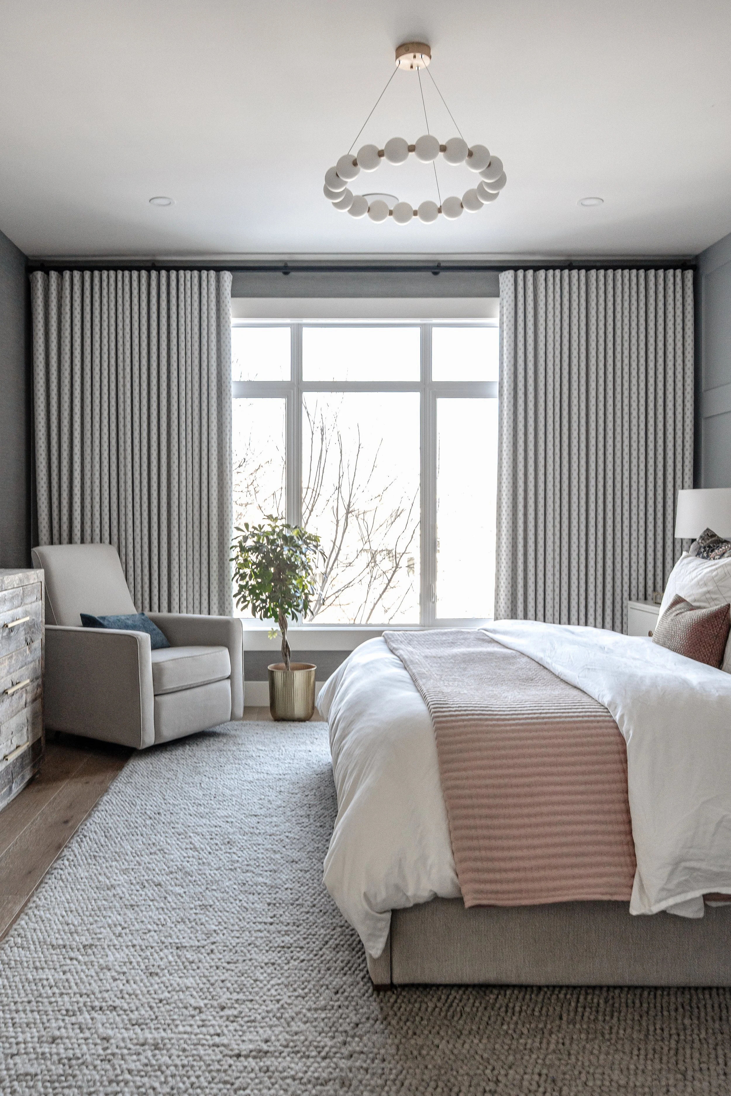Scenic bedroom with large window, white curtains, beige bed, Armchair, plant, circular chandelier, beige rug, natural light.