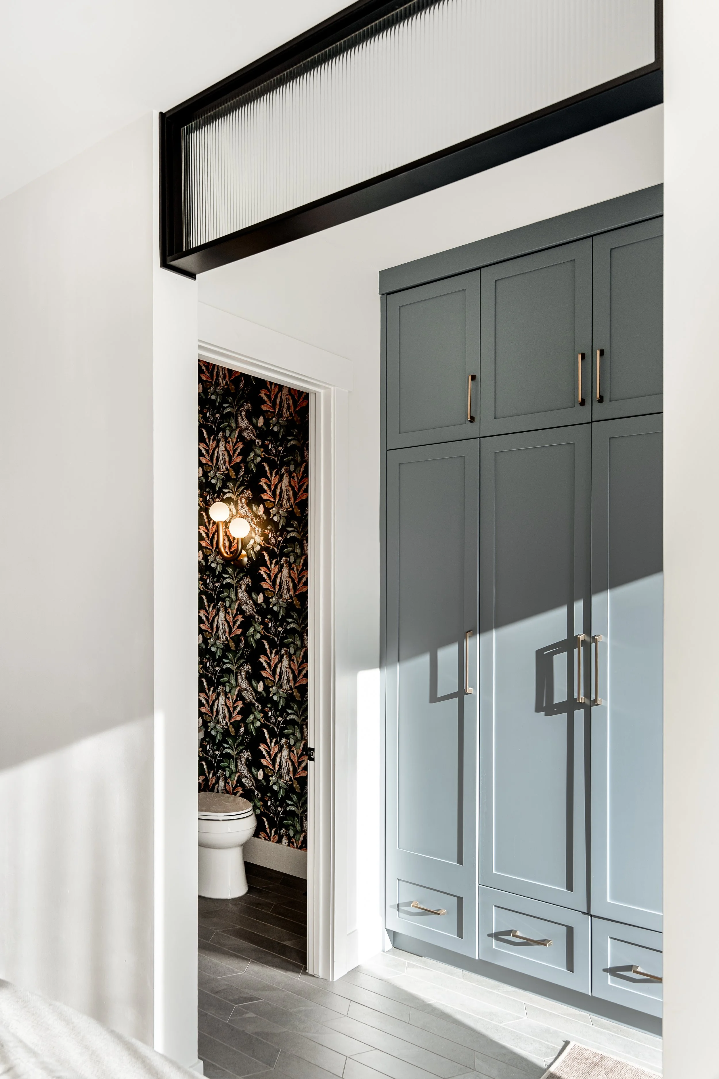 View of a modern bathroom with gray cabinetry, a small toilet room with floral wallpaper, and a white sink with a round mirror above it.