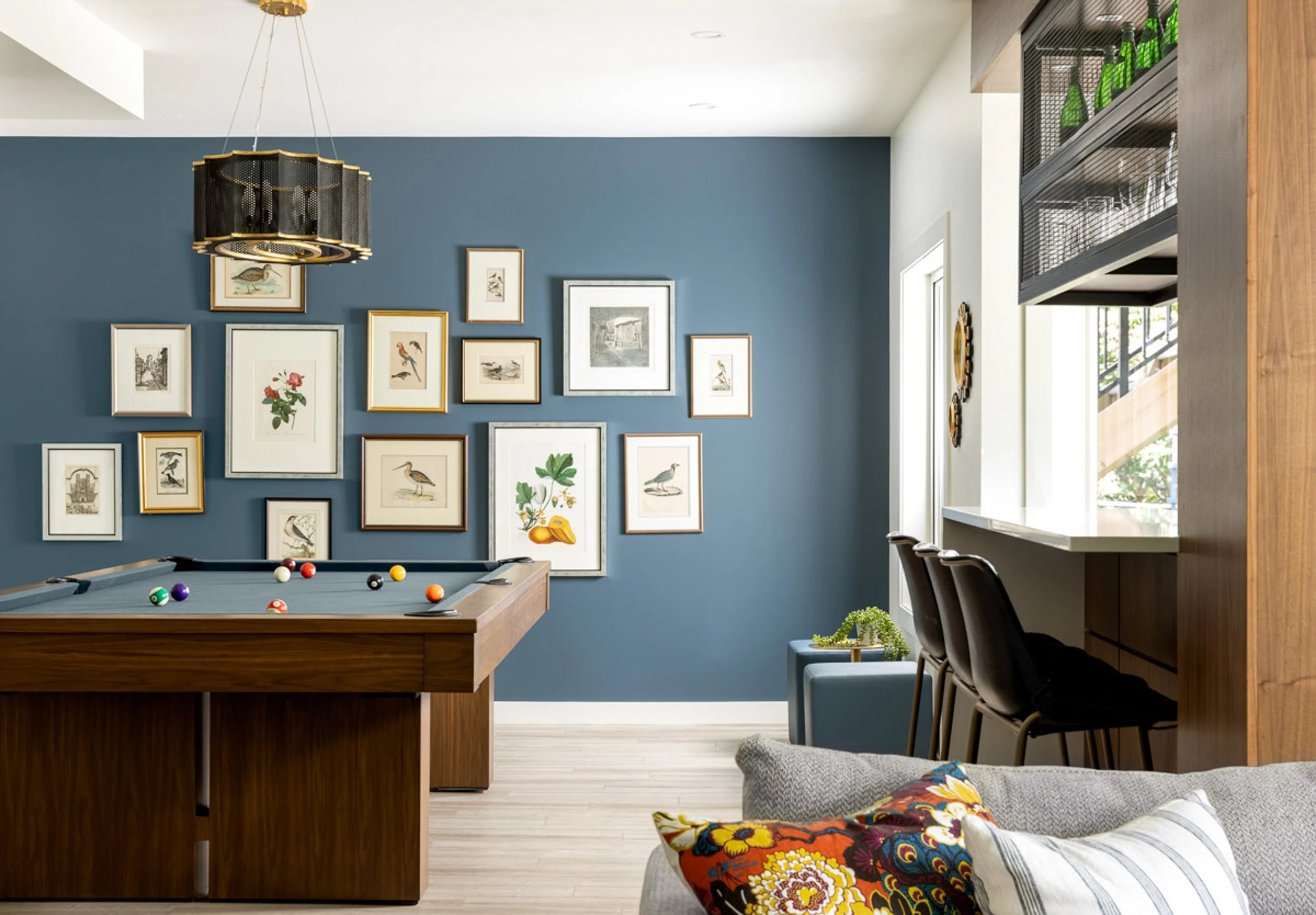Modern game room with a pool table, blue accent wall featuring framed artwork, contemporary chandelier, bar area with stools, and a couch with decorative pillows.