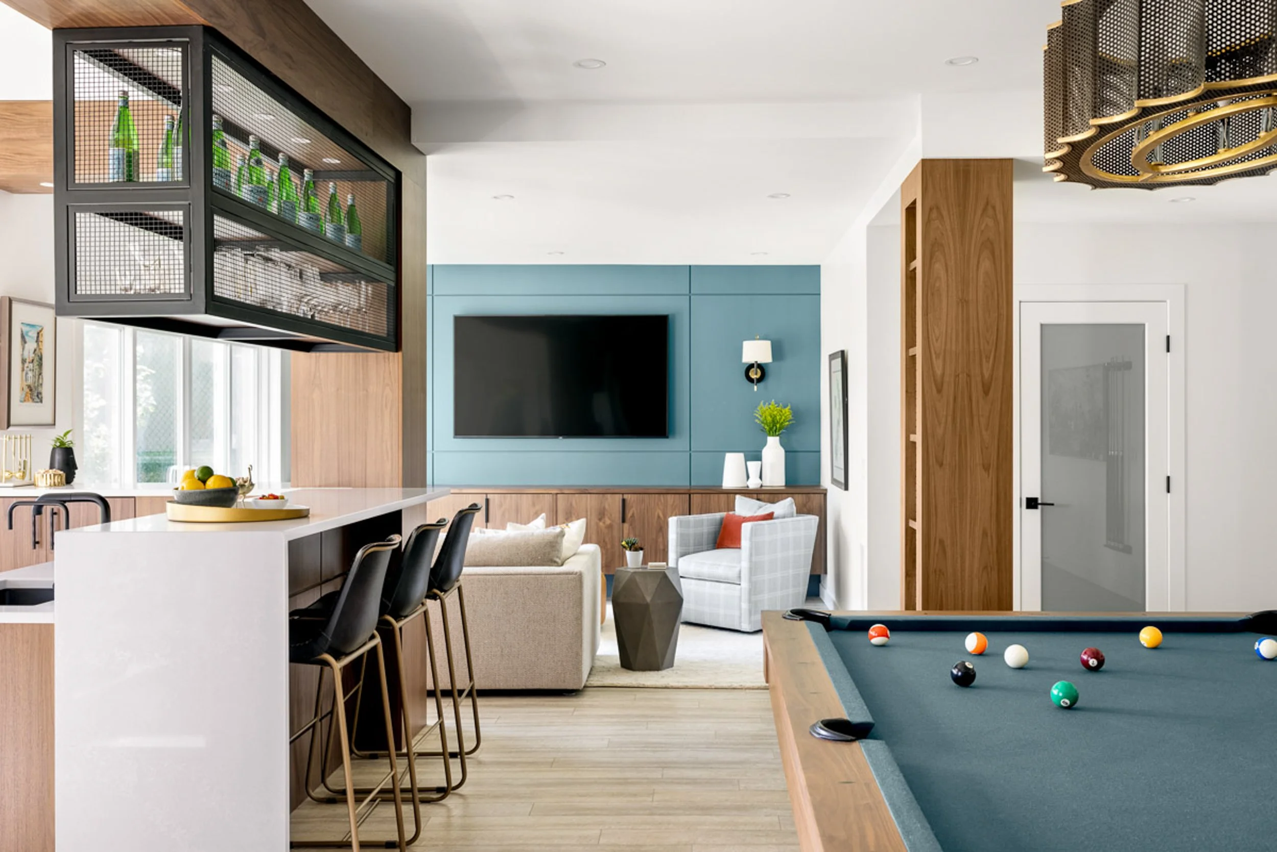 Modern game room with pool table, bar counter, bar stools, sofa, TV, and wooden accents.