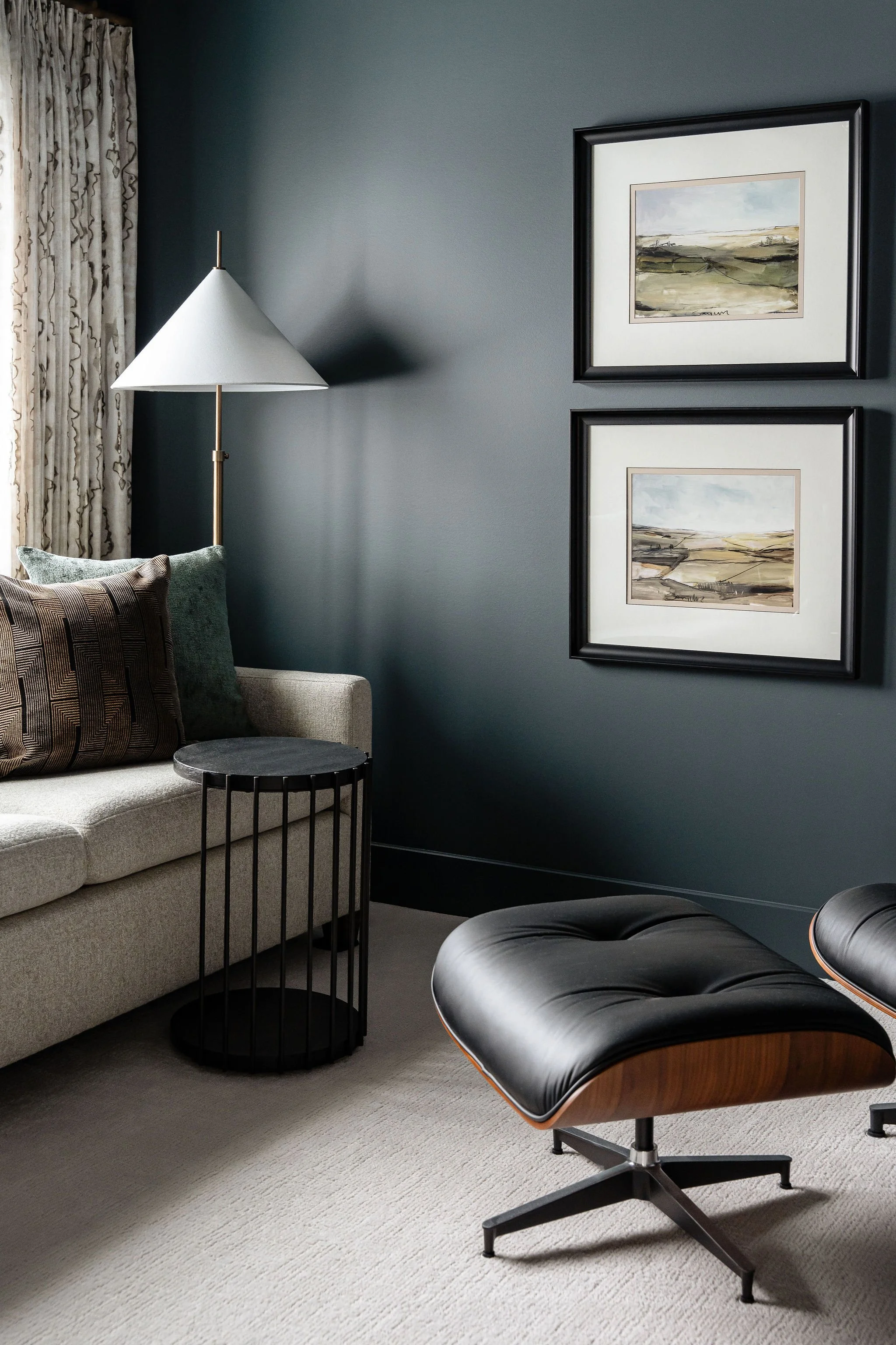 Cozy living room with a beige sofa, black wire side table, black leather lounge chair, a floor lamp with a white shade, gray carpet, and framed landscape artwork on a dark gray wall.