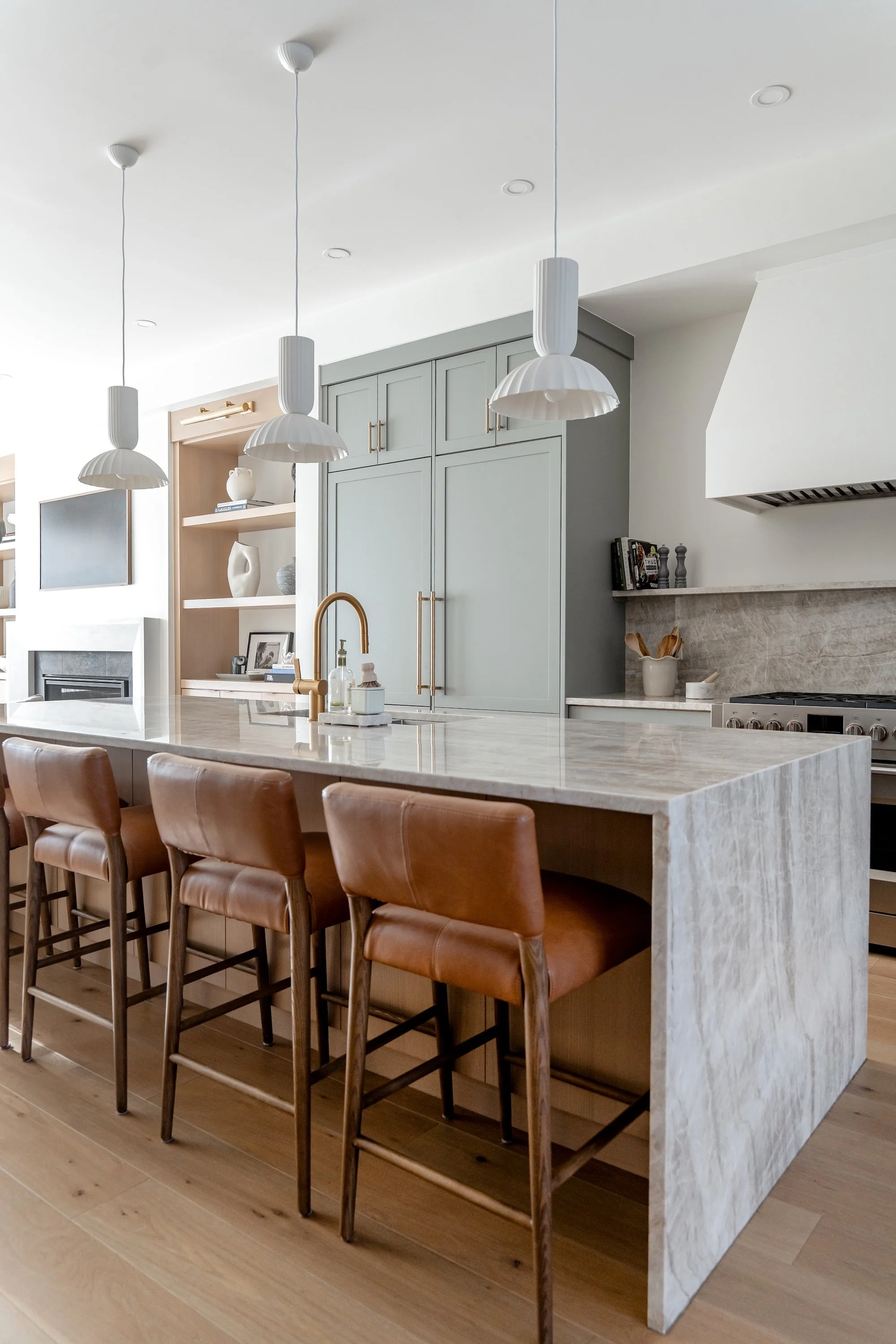 Kitchen island with a marble top, four brown leather barstools, white ceiling with three hanging pendant lights, gray cabinets, and a stove near the wall.