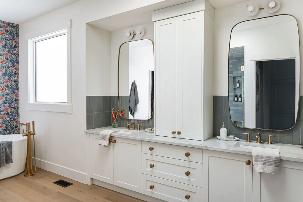 Bright bathroom with a white vanity, two oval mirrors, gold fixtures, a bathtub with a gray towel, a window, and floral wallpaper on one wall.