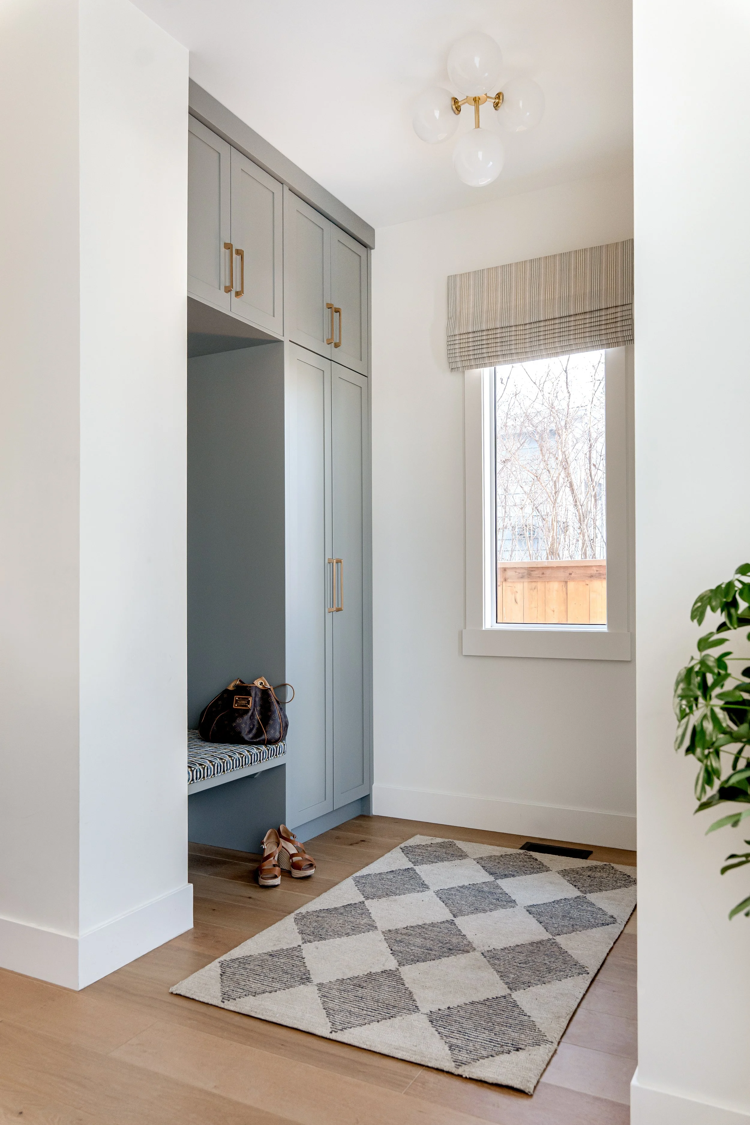 A cozy entryway with a window brightening the space, light gray cabinets, a bench with a patterned cushion, shoes on the floor, and a geometric rug. A handbag is on the bench, and a plant is partially visible on the right.