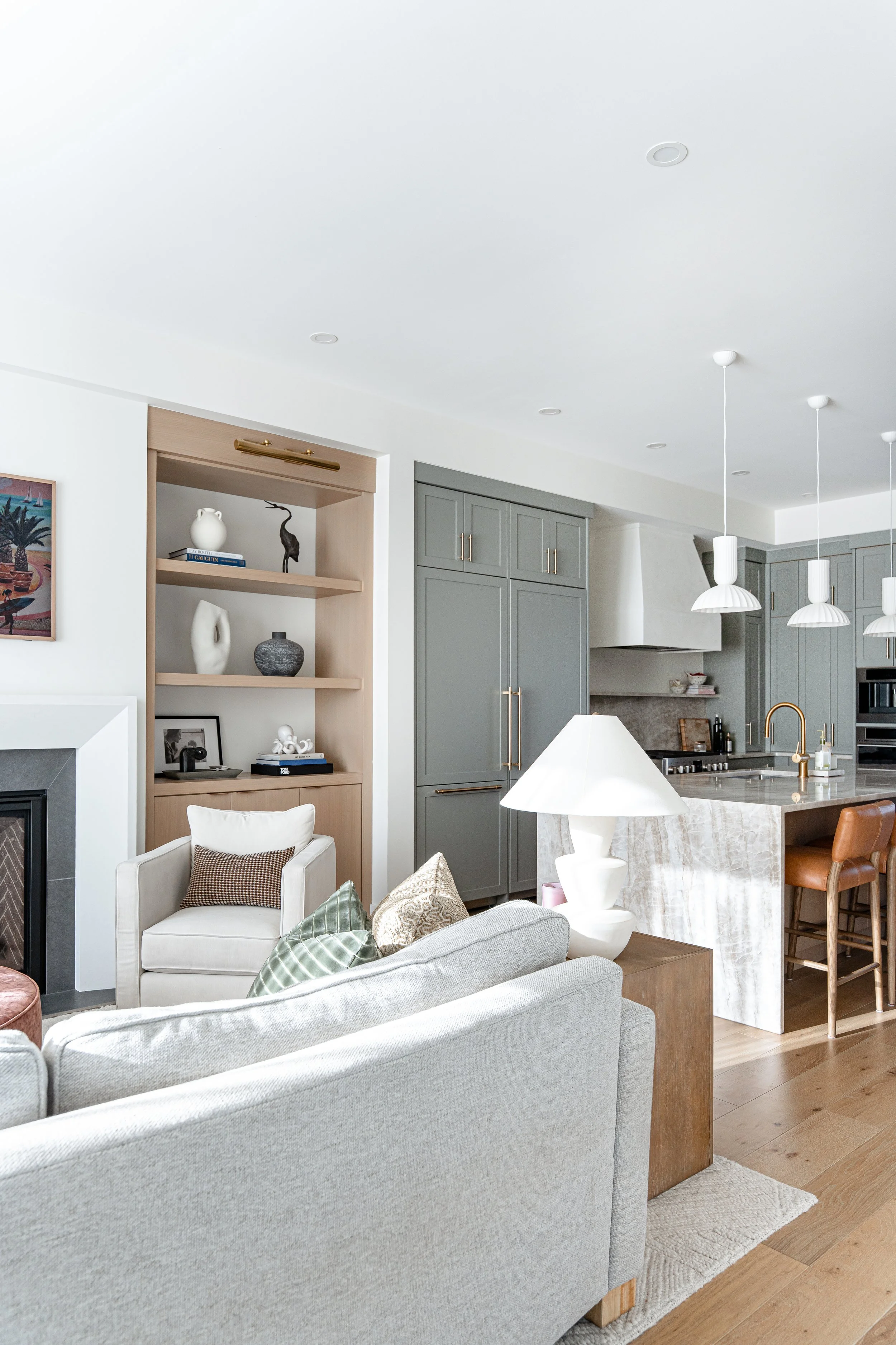 Interior view of a modern living room with a light gray sofa, a white armchair with a brown pillow, a wooden side table with a white lamp, and view into a kitchen with gray cabinets and hanging white pendant lights.