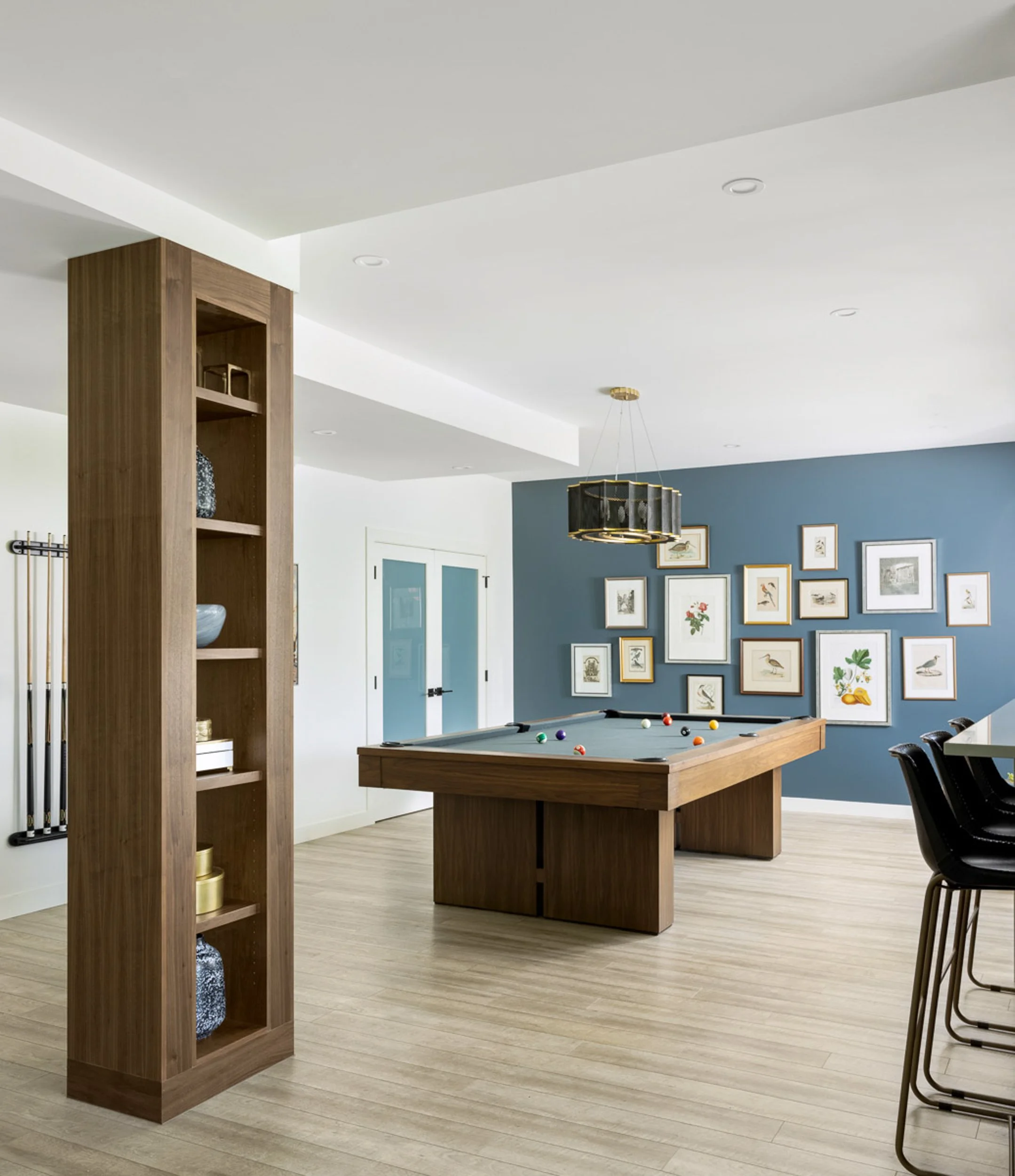 Modern game room with a pool table, wooden shelving, wall art, and blue accent wall. Features a wooden floor, pool cues on the wall, and a chandelier above the table.