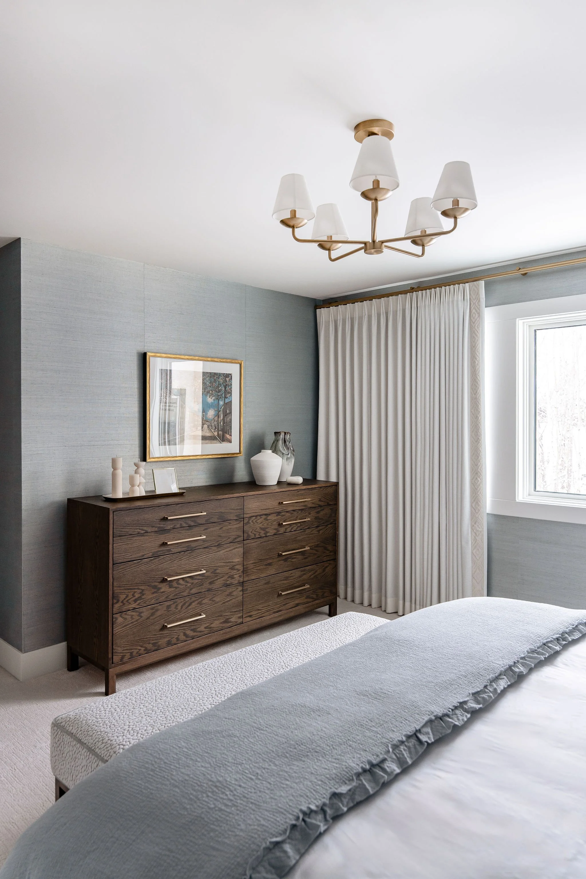A bedroom corner with a wooden dresser, framed artwork, decorative vases, white curtains, a window, and a chandelier on a white ceiling.