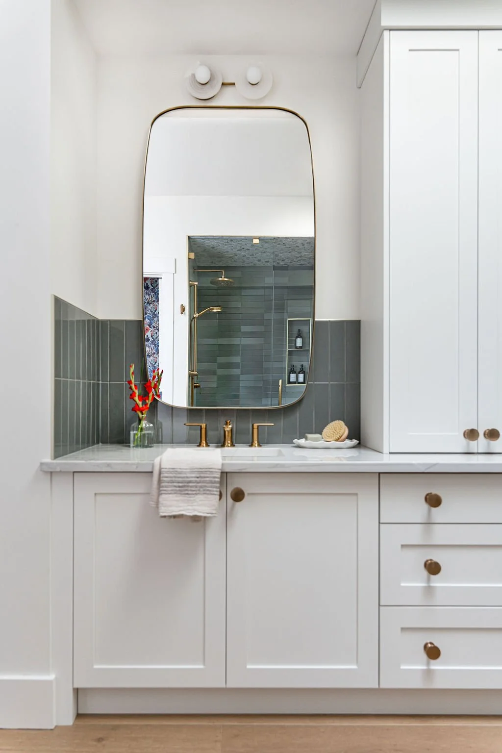 A bathroom vanity with a large mirror, white cabinetry with brown knobs, a white countertop, gold fixtures, and a small vase with red flowers. Visible shower area with a gold showerhead and gray tiled walls reflected in the mirror.