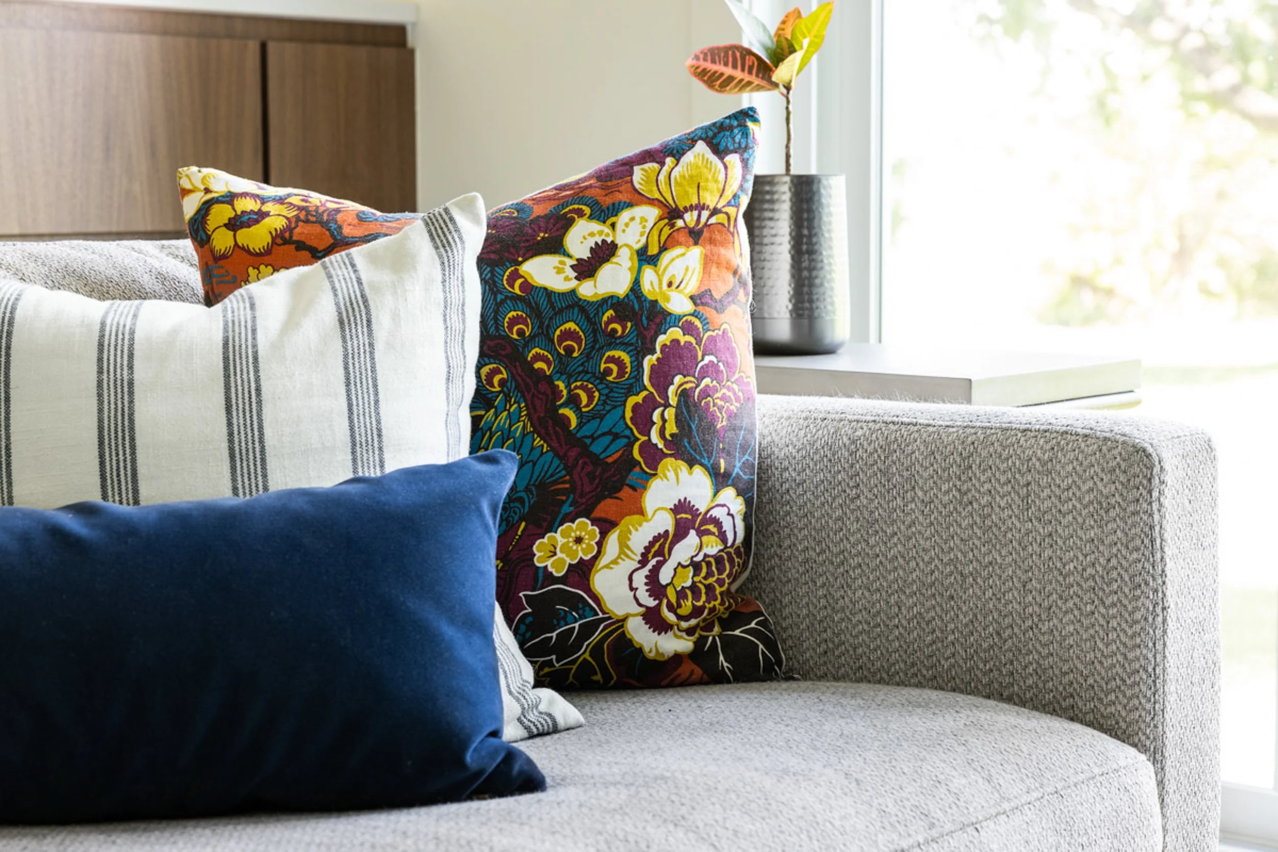 Living room with a modern gray sofa adorned with decorative pillows featuring floral and striped patterns, and a blue solid pillow. A small potted plant and a book are placed on the armrest near a window.