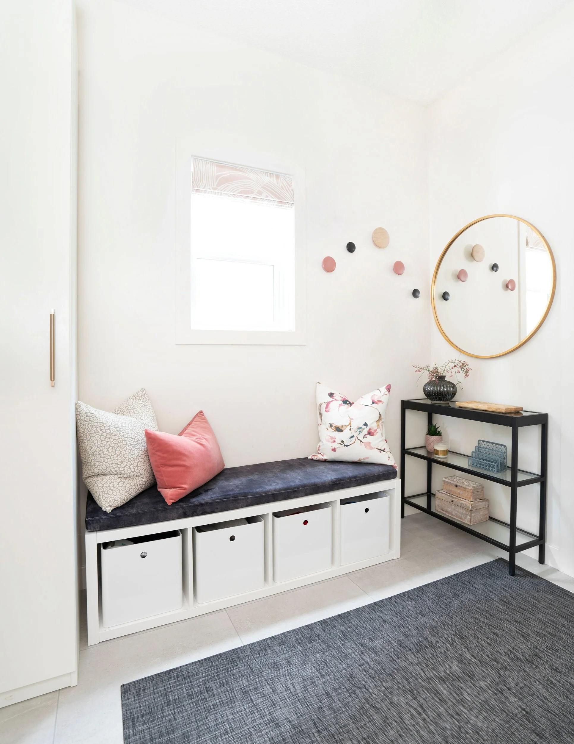 Modern entryway with a cozy bench covered in cushions, surrounded by storage cubes below. A small black metal shelf holds decorative items next to a round mirror on the wall. The window lets in natural light, and a gray textured rug covers the floor.