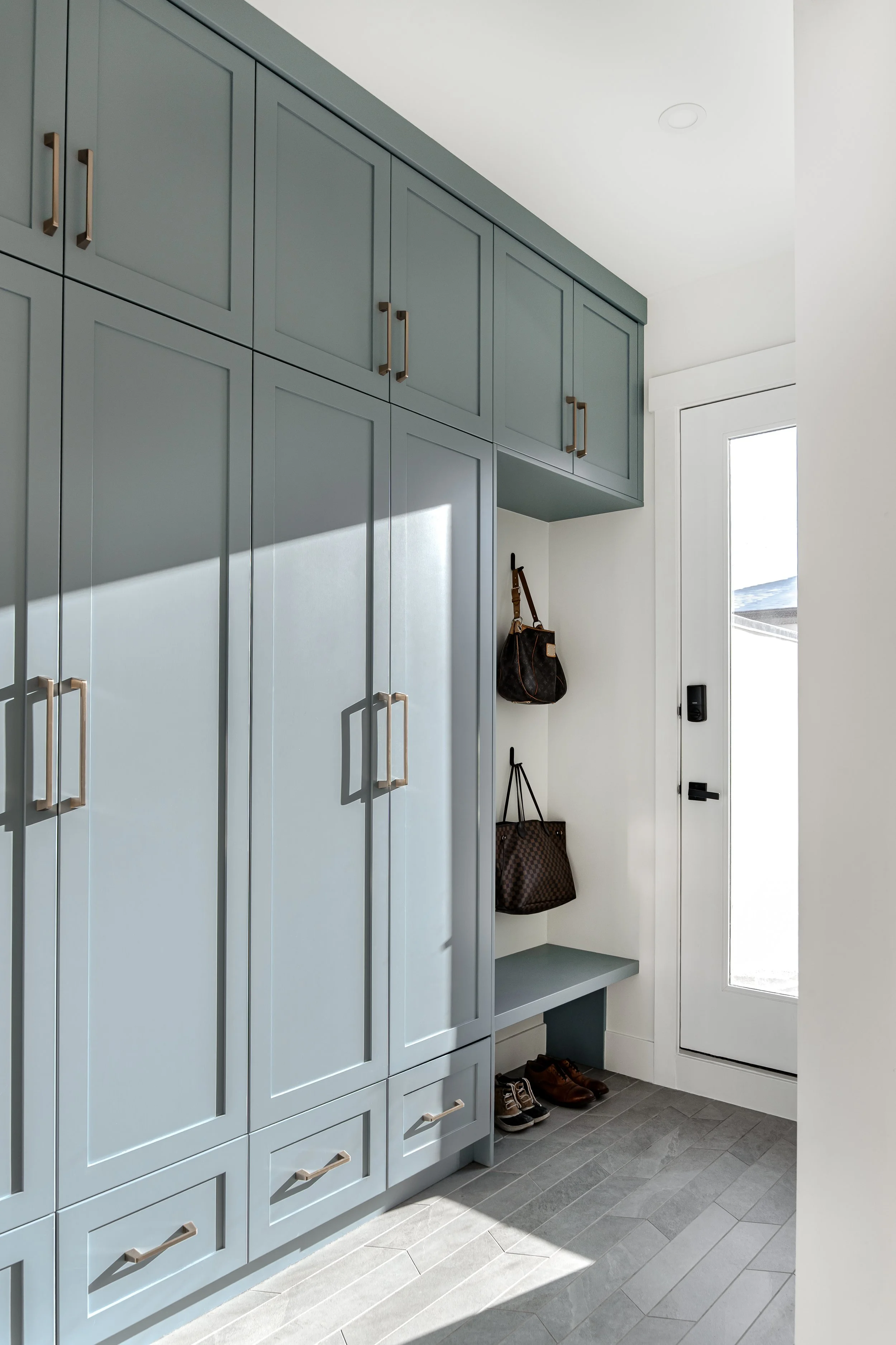 Light blue mudroom with tall cabinets, a small bench, handbags hanging, and shoes on the floor near a white door with glass panel.