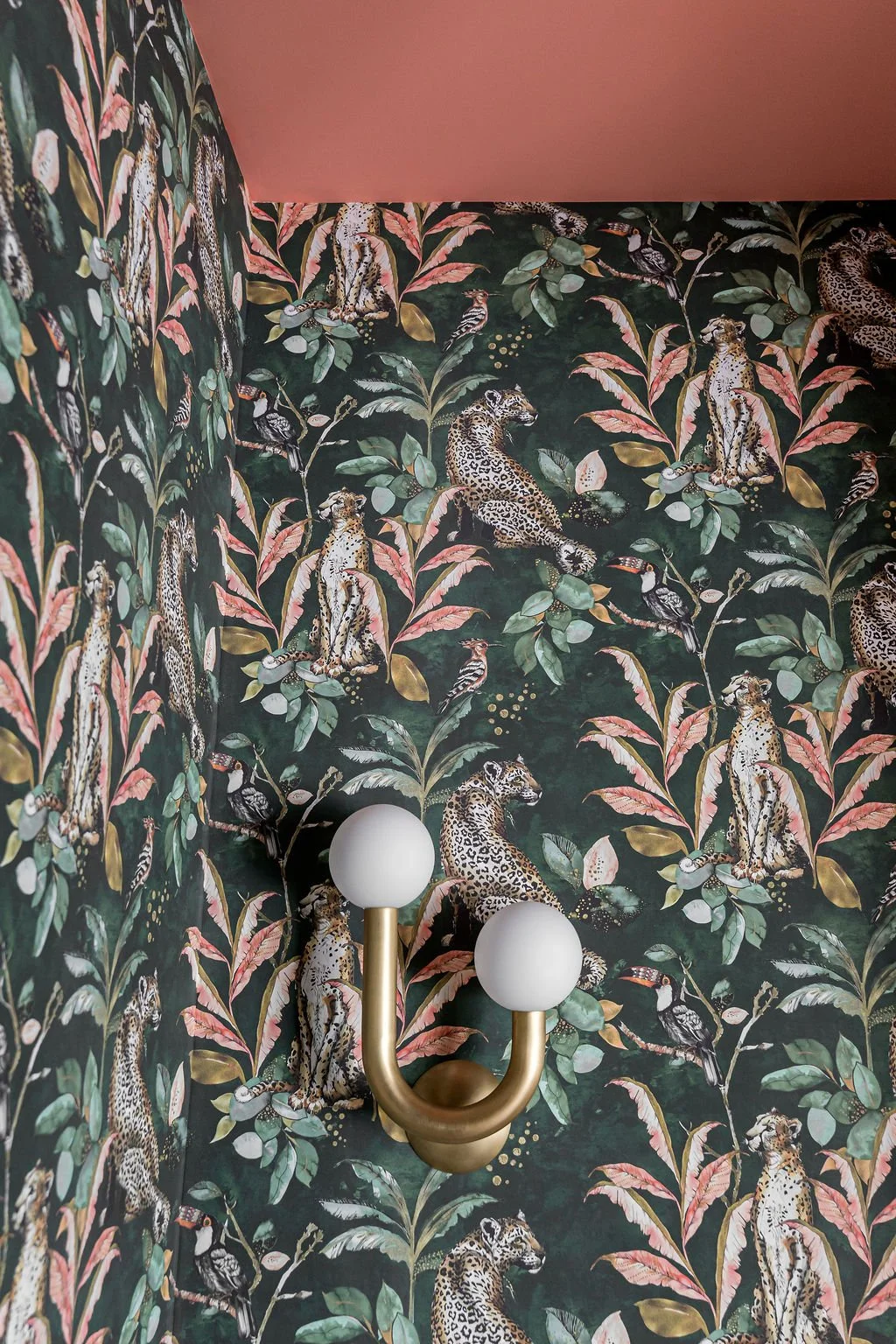 A corner of a room with wallpaper featuring a jungle theme, with jaguars, tigers, and tropical leaves, and a gold sconce with two white globe lights.