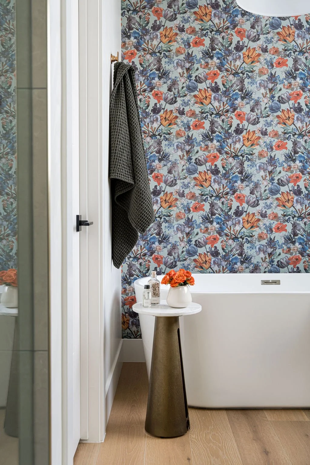 Bathroom with floral wallpaper, a white bathtub, a small round white table holding a white vase with orange roses, and toiletries including a soap dispenser and lotion bottle. A dark towel hangs on a hook on the white wall.