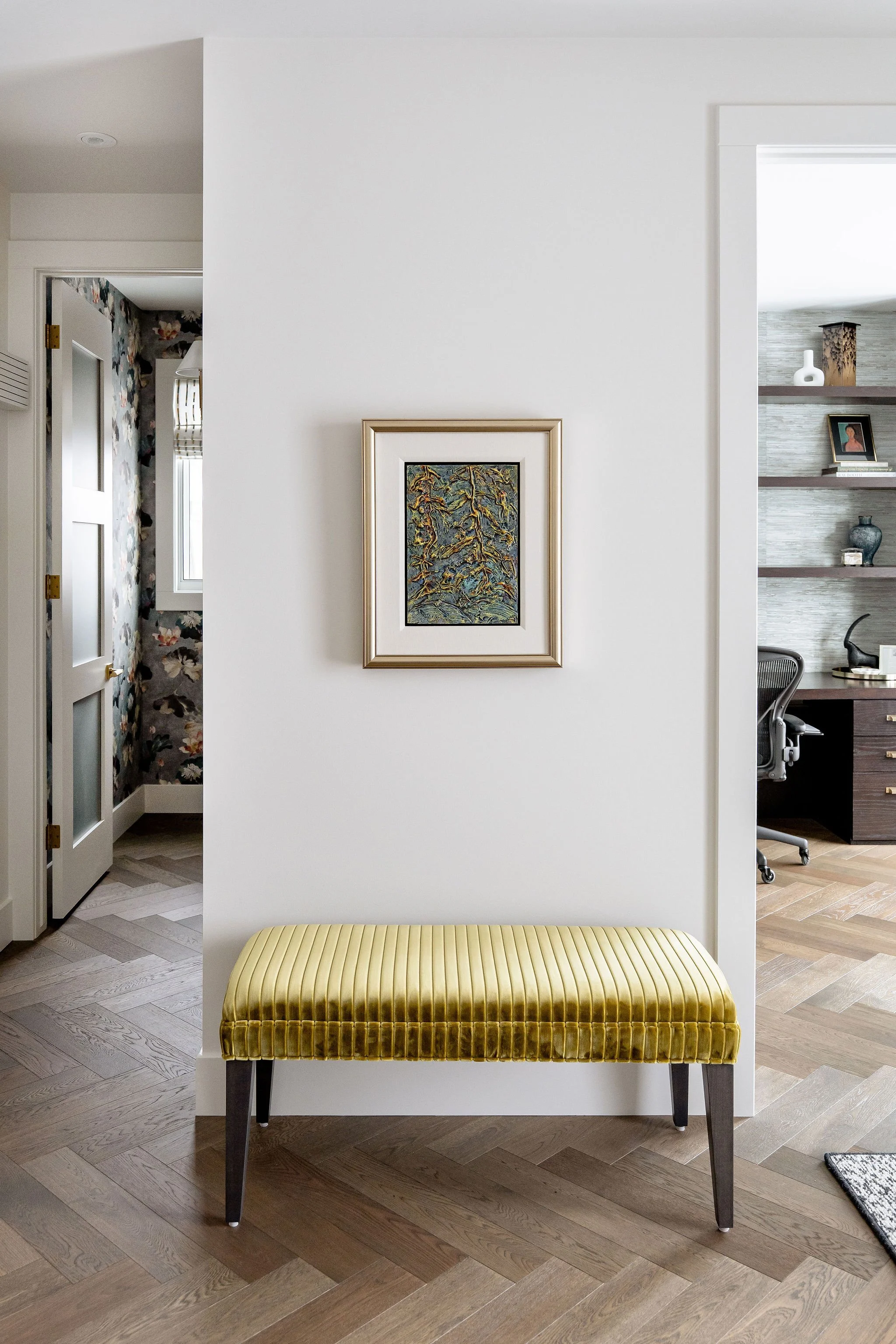 A hallway with a patterned wallpaper on the left and a room visible through a doorway on the right. A framed painting hangs on a white wall. In front of the wall, there is a yellow velvet bench with dark wooden legs on a light wood herringbone floor.