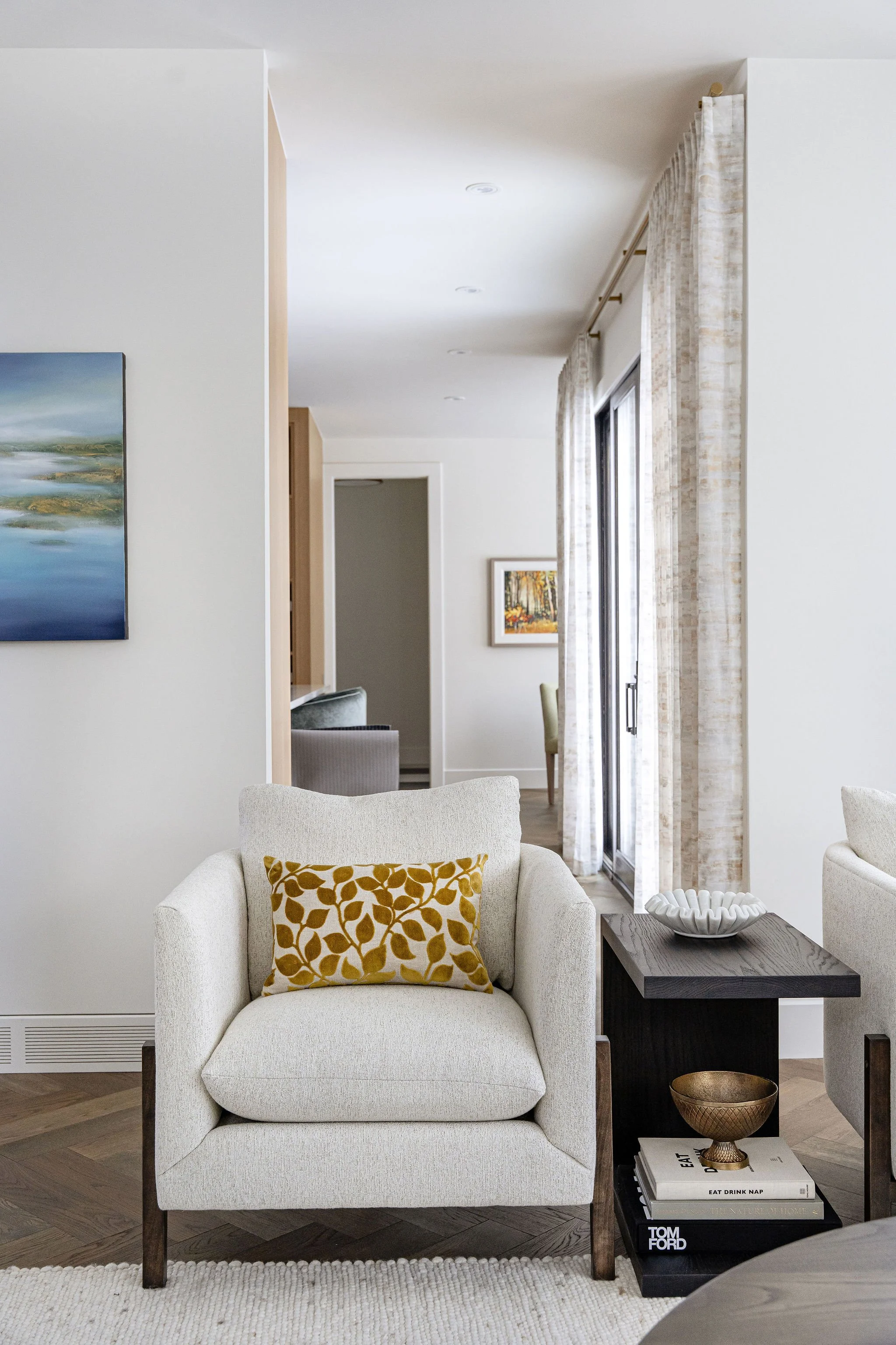 A living room with a beige armchair that has a yellow and brown patterned pillow, a dark wooden side table with books and a decorative bowl, curved beige curtains, and a large window. There are paintings on the walls, and the floor is hardwood with a