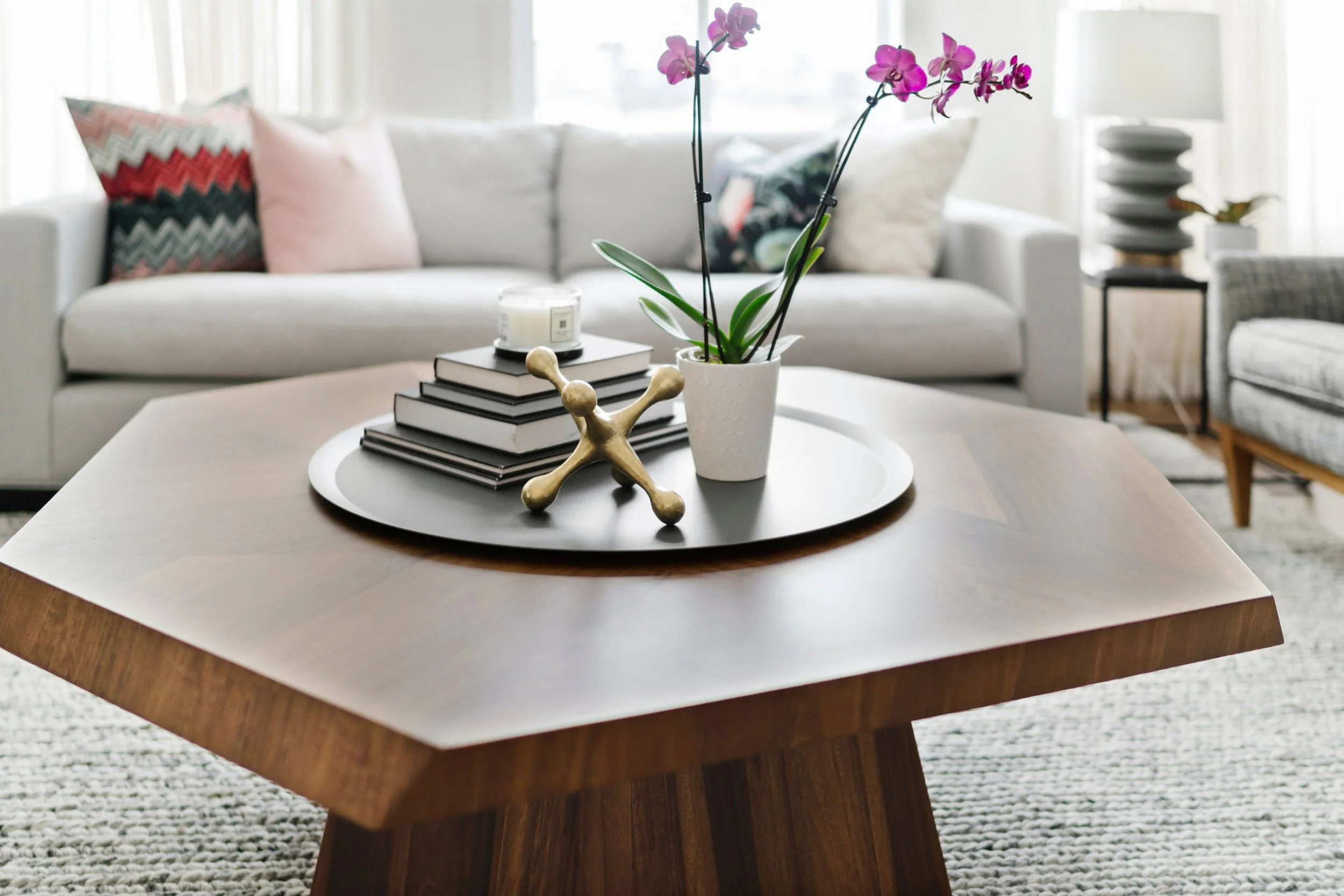 Modern living room with hexagonal wooden coffee table, decorative tray, potted orchid, books, gold sculpture, and sofa with pillows.