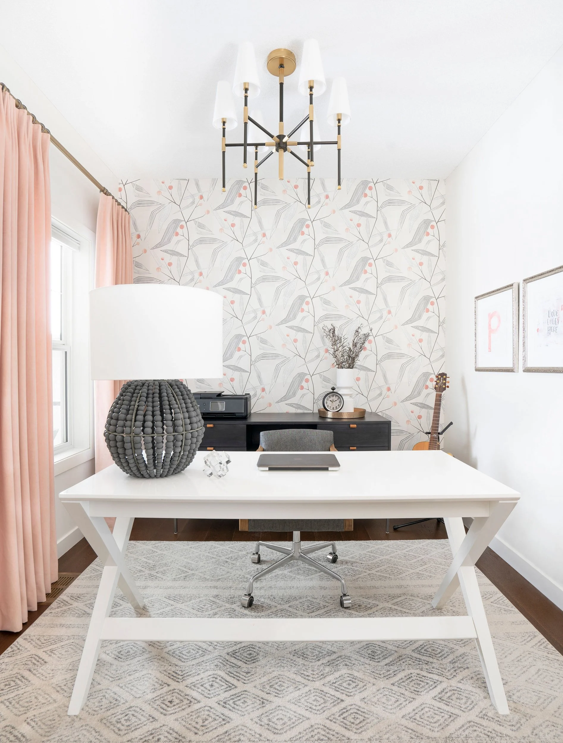 Modern home office with a white desk, a black woven lamp, and a chair on a patterned rug. The room has floral wallpaper, a ceiling light fixture, and pink curtains. A guitar and decorative items are on a black cabinet against the wall.