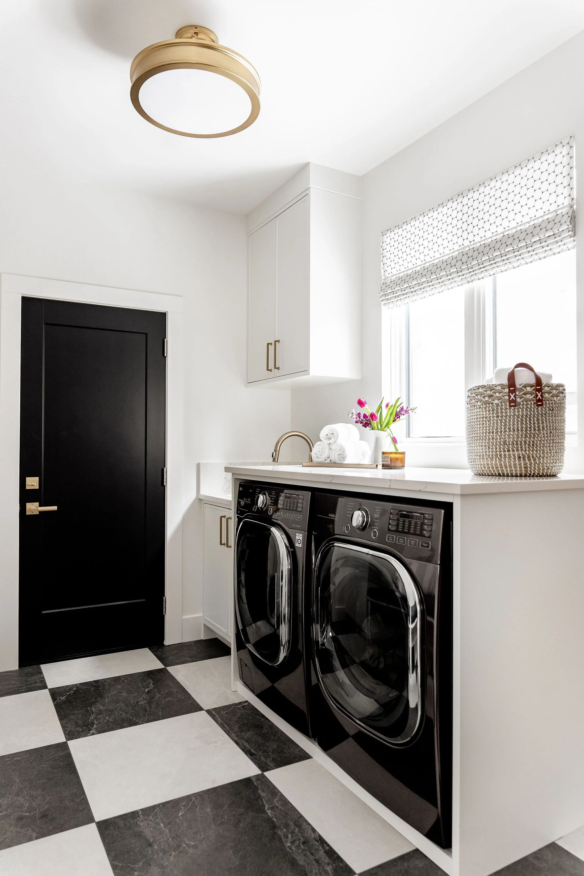 A laundry room with a black front-load washer and dryer set, white countertops, a window with a patterned Roman shade, and a shelf with decorative items including folded towels, a vase of flowers, and a woven basket.
