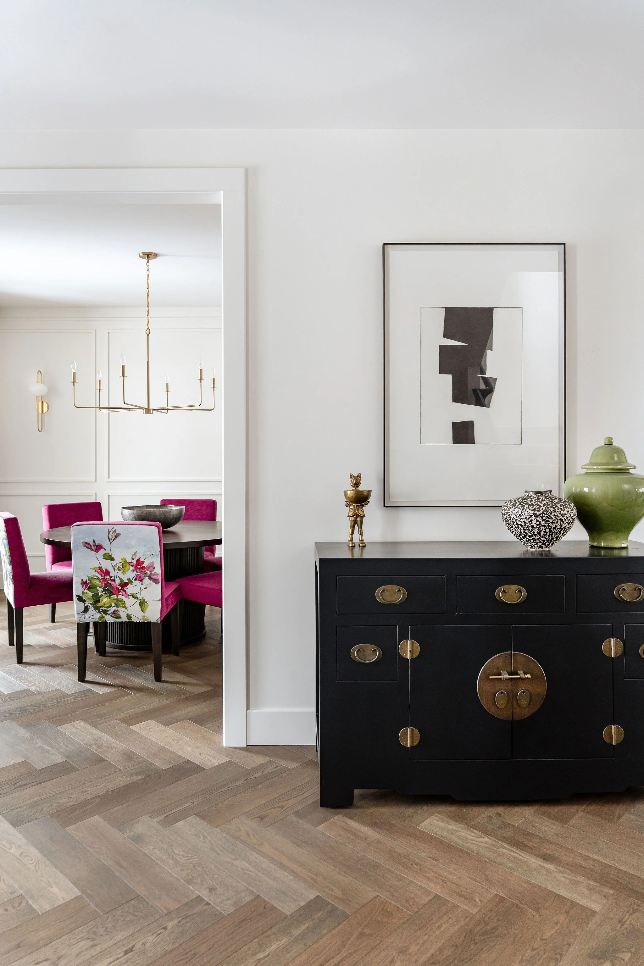 A dining room with a black sideboard decorated with vases and a sculpture, a white wall with a framed abstract artwork, and a table with pink chairs and a chandelier visible in an adjoining room.