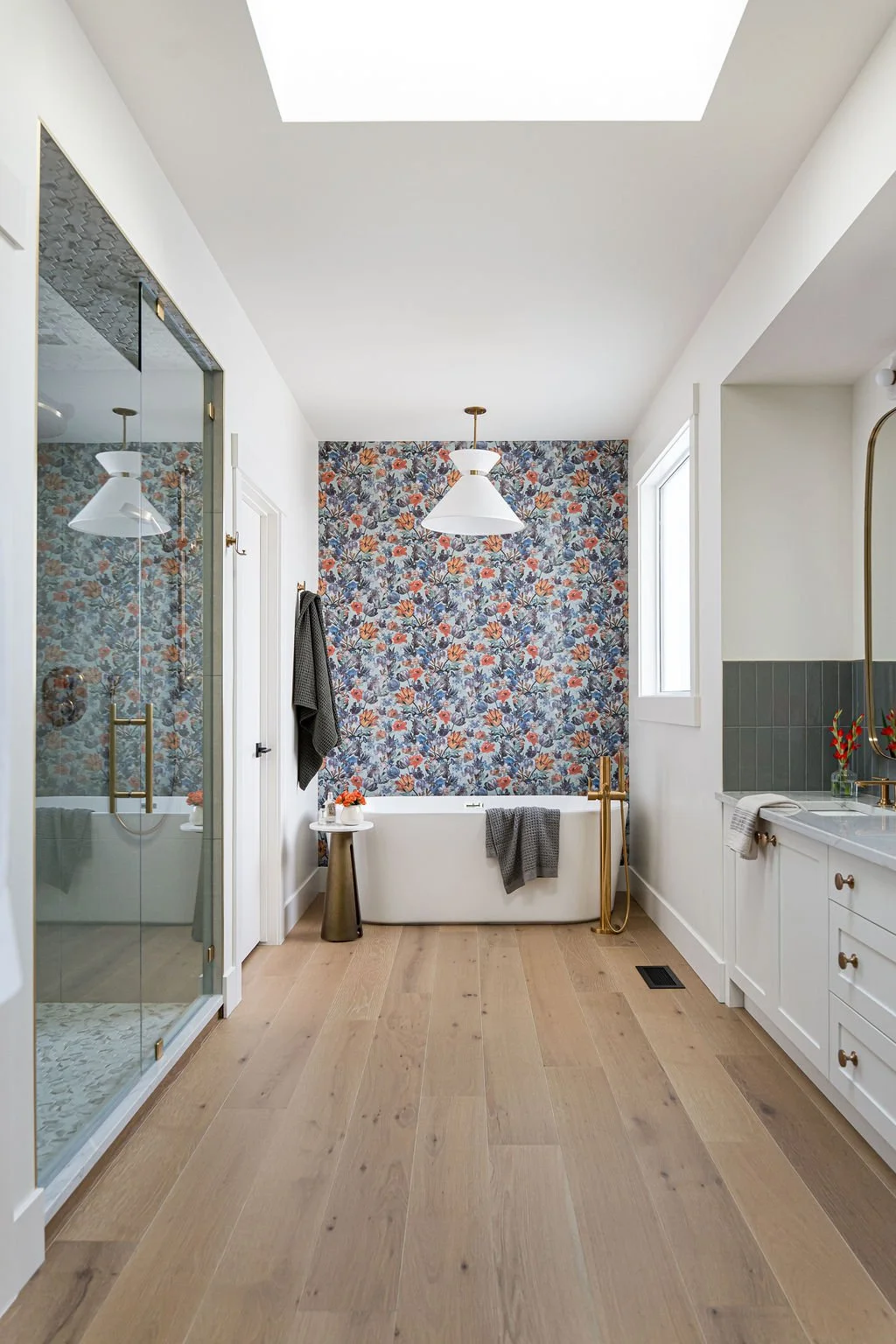 Modern bathroom featuring a skylight, floral accent wall behind a white bathtub, a separate glass-enclosed shower, a white vanity with gold fixtures, and wooden flooring.