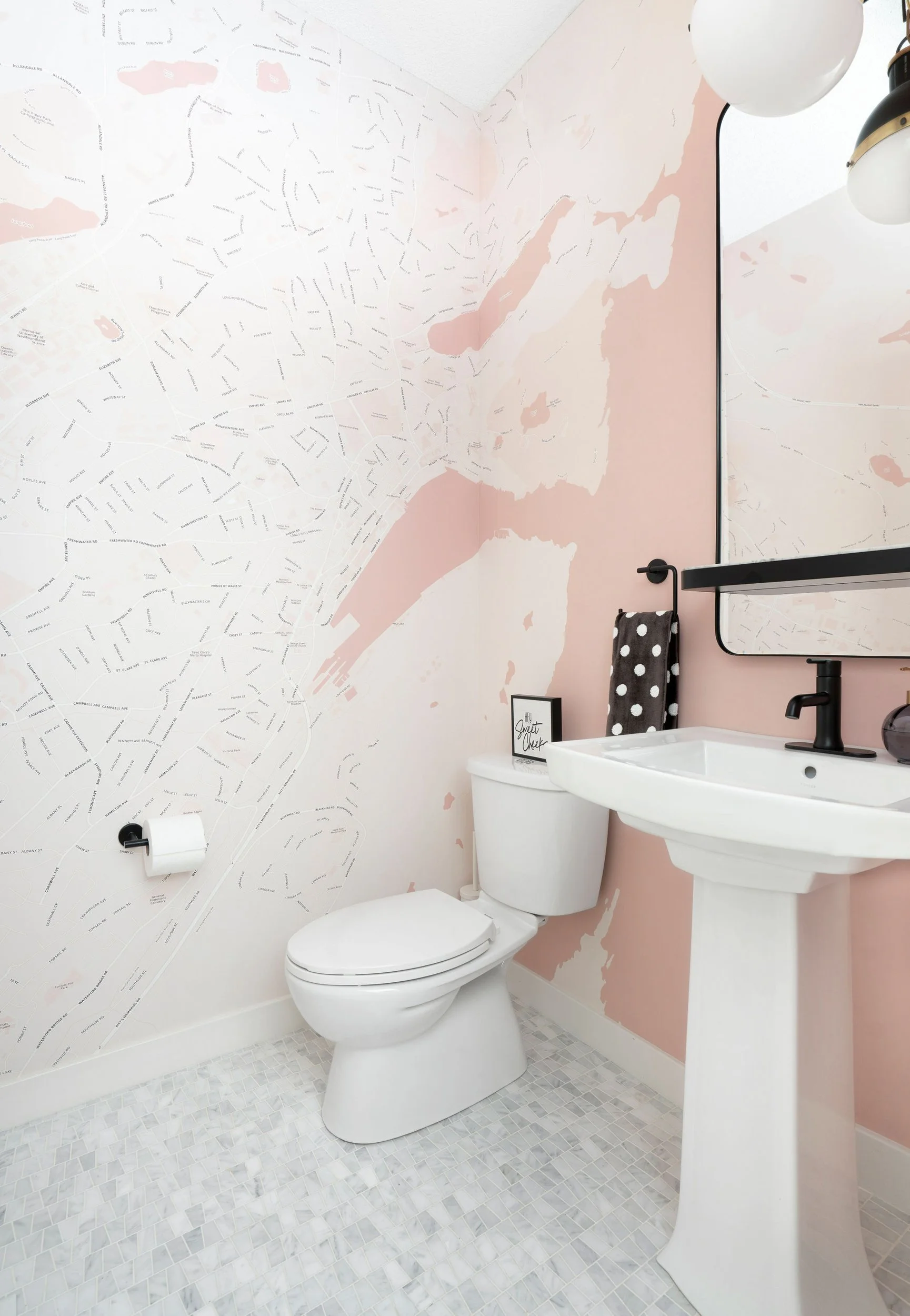 A modern bathroom with a map-themed wallpaper, featuring a white toilet and pedestal sink, black faucet, and a black framed mirror. The floor is tiled, and there is a black polka dot towel and a small framed sign on the toilet tank.