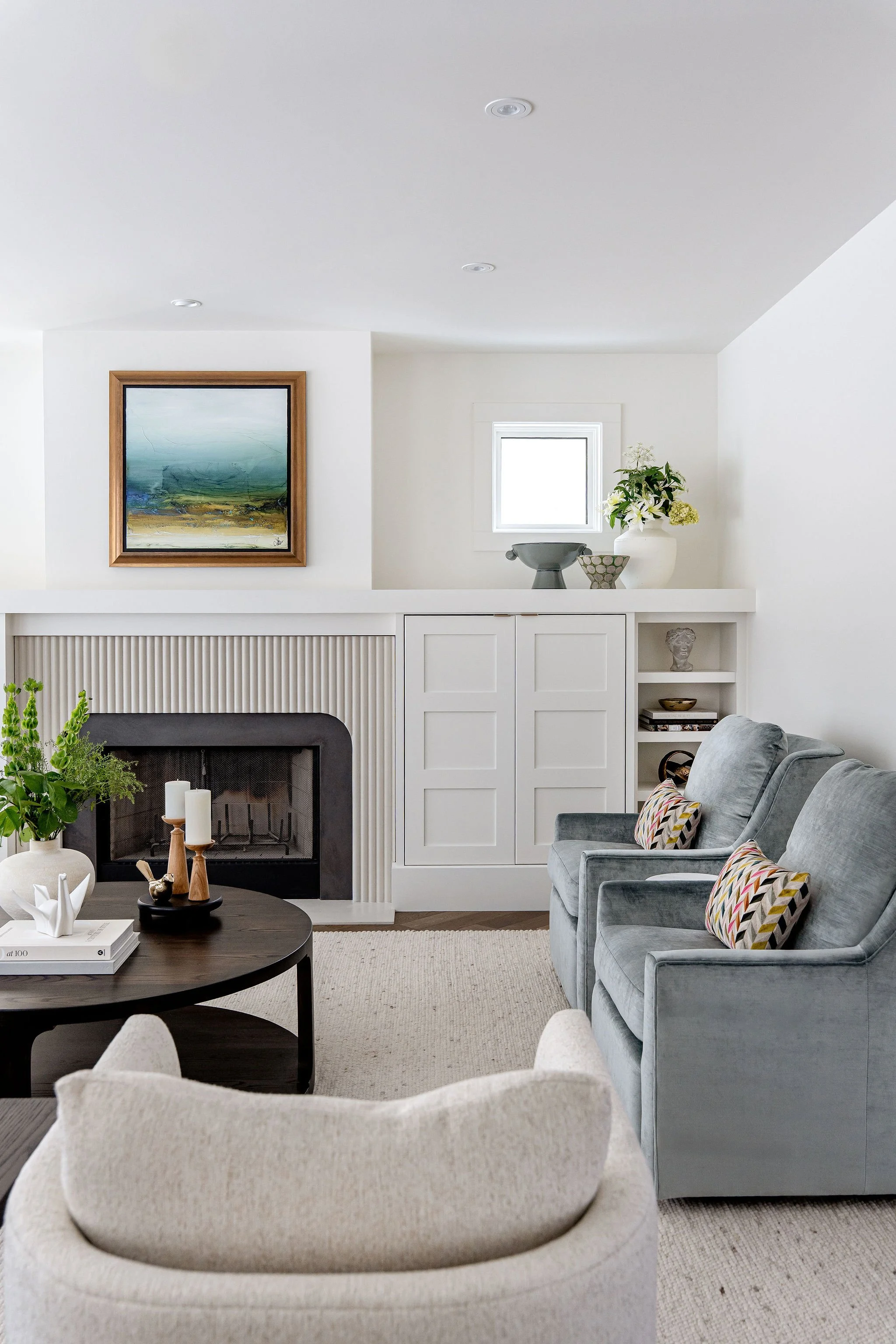 Living room with a white built-in cabinet, a fireplace, and a gray armchair. Decor includes a table with candles, a white sculpture, green plants, and framed artwork on the wall.