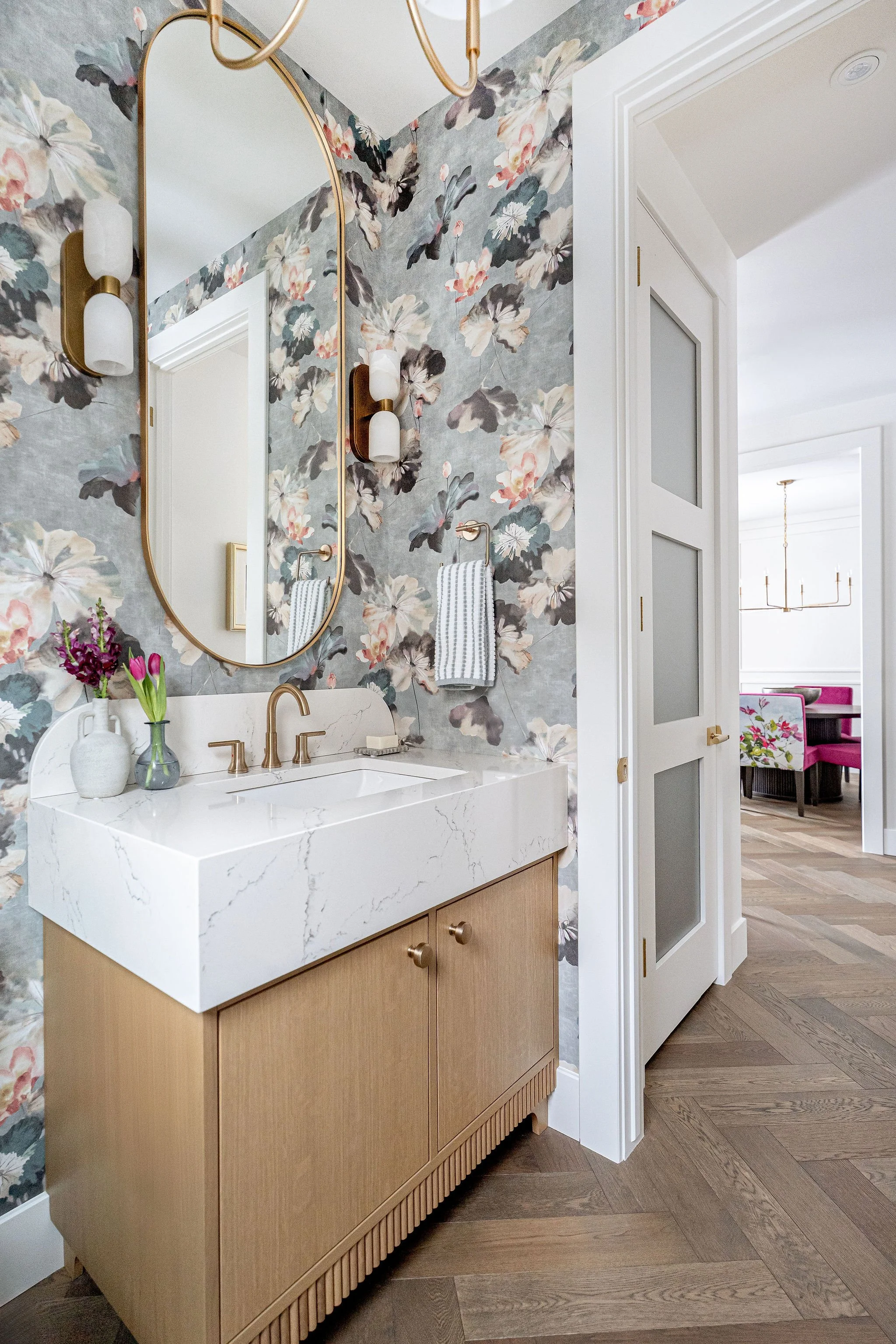 A bathroom vanity with a white marble countertop, brass faucet, and a light wood cabinet below with decorative trim. Behind the vanity is a wall with floral wallpaper featuring large, soft-colored flowers. To the right, an open white door with glass 