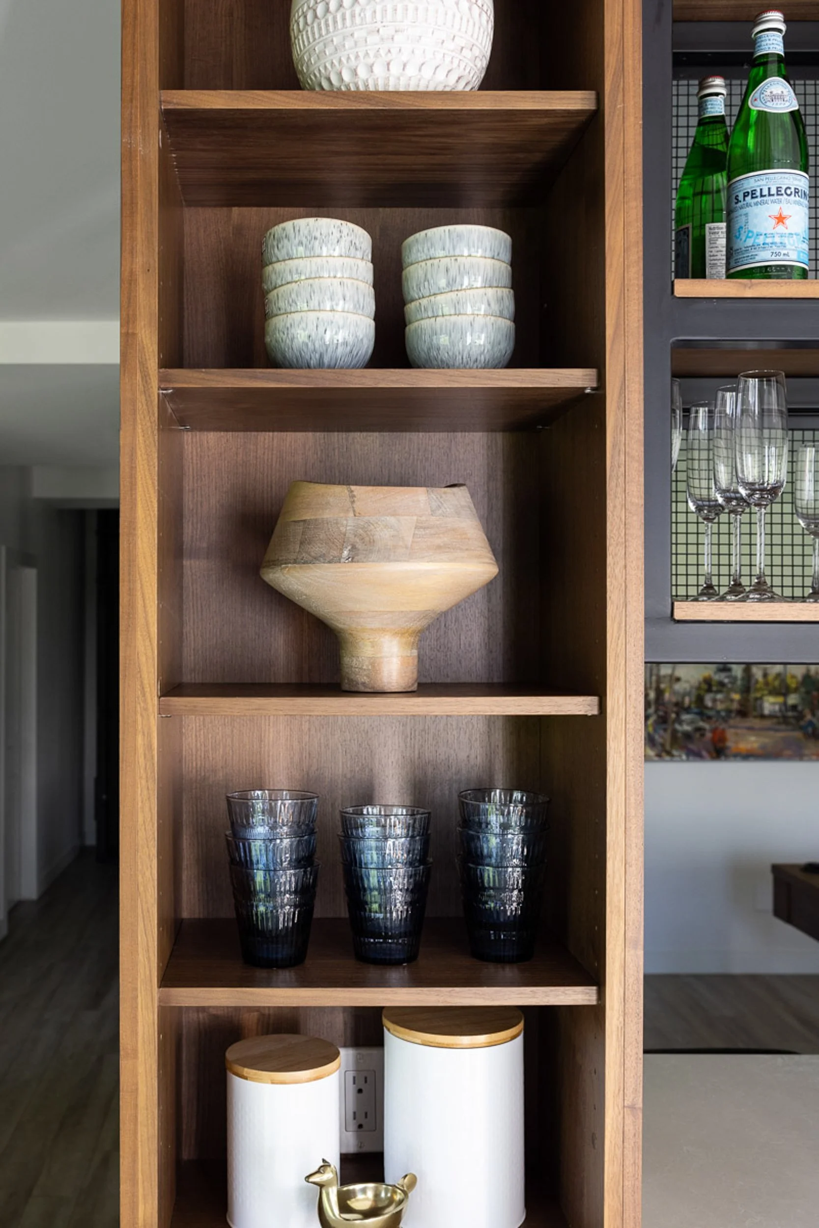 A wooden cabinet with shelves displaying decorative bowls, glasses, a sculpted vase, ceramic canisters with wooden lids, and a small golden figurine. To the side are glassware and bottled water.