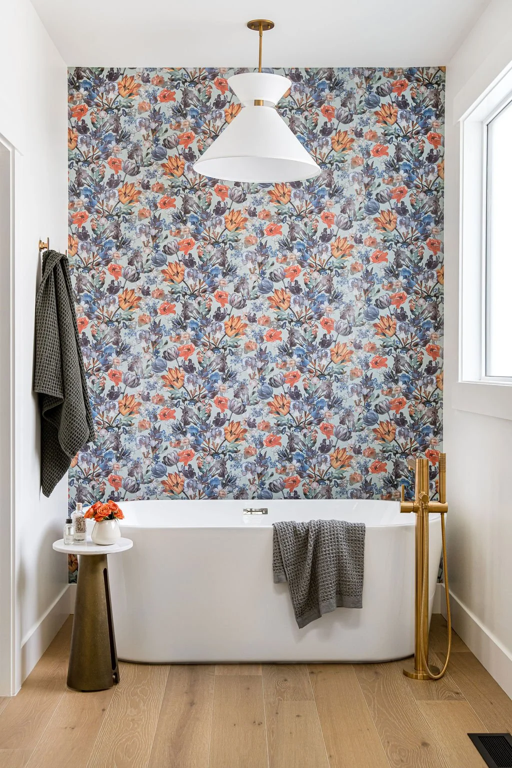 Bathroom with a white bathtub, floral wallpaper, a small side table with decorative items, and a gold bathtub faucet with a handheld shower.