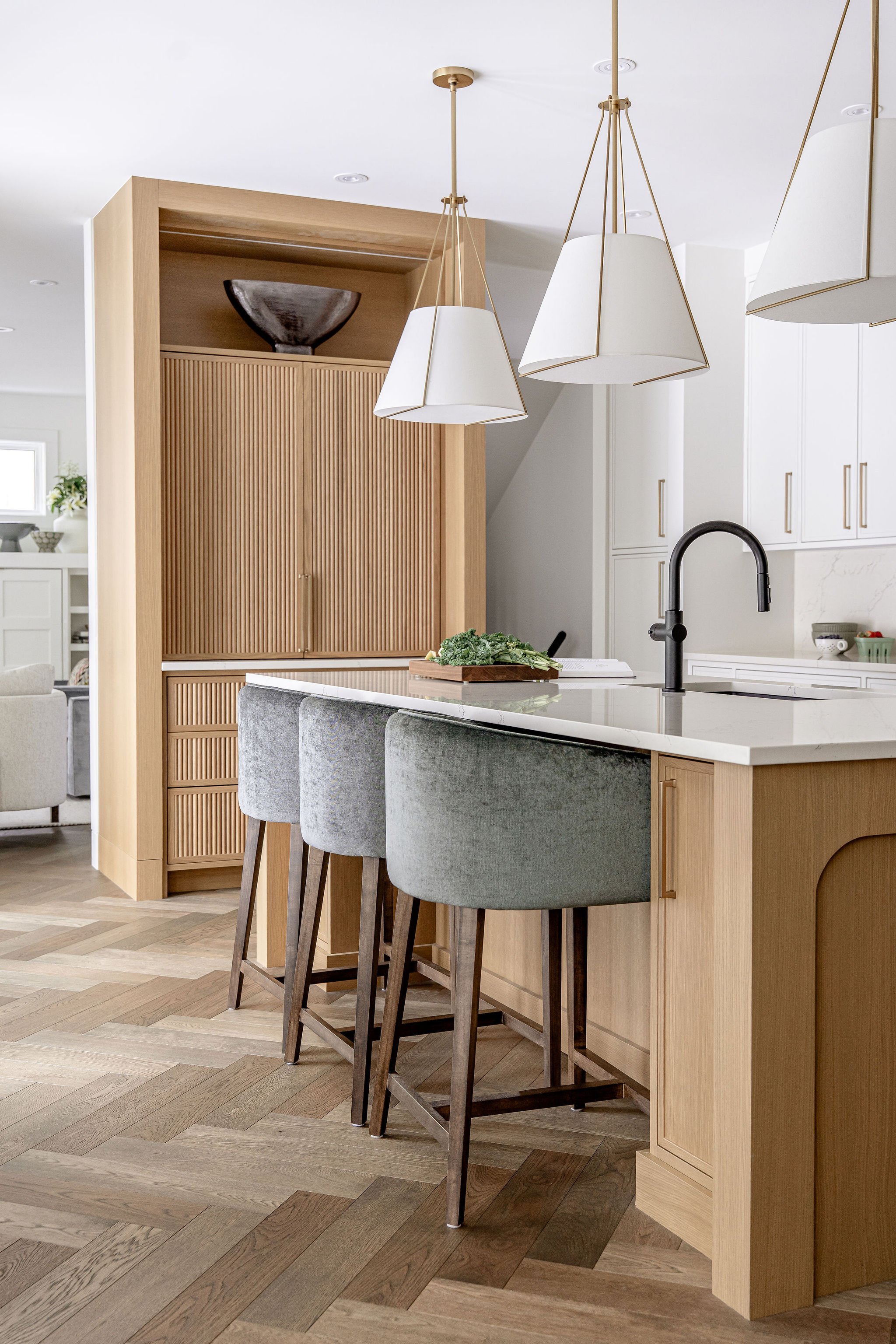Modern kitchen with light wood cabinets, a white countertop, black faucet, four upholstered gray bar stools, and a pendant light fixture with three white shades.