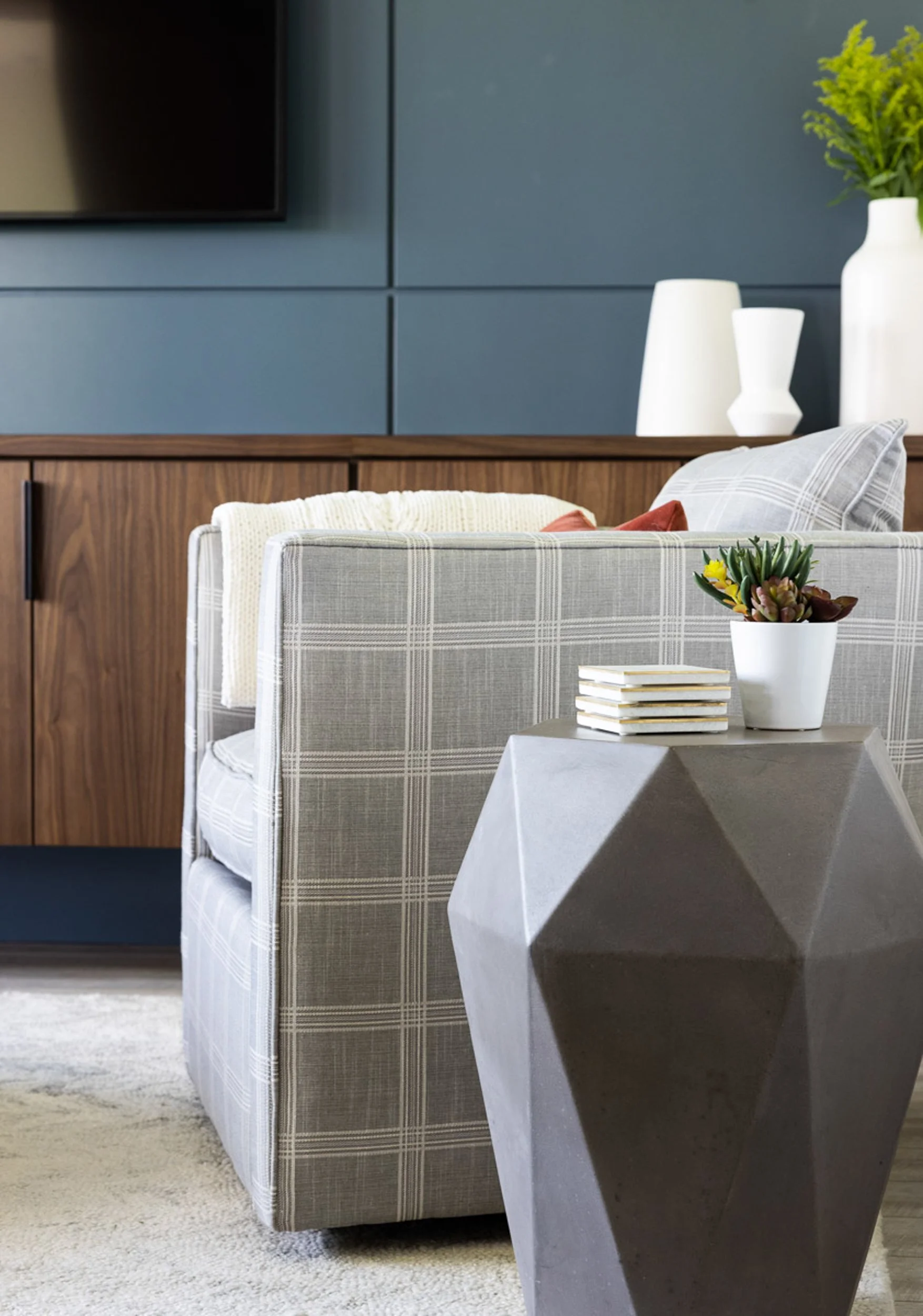 Modern living room with geometric gray side table, gray plaid armchair, wooden cabinet, decorative white vases, and a small potted plant with coasters.