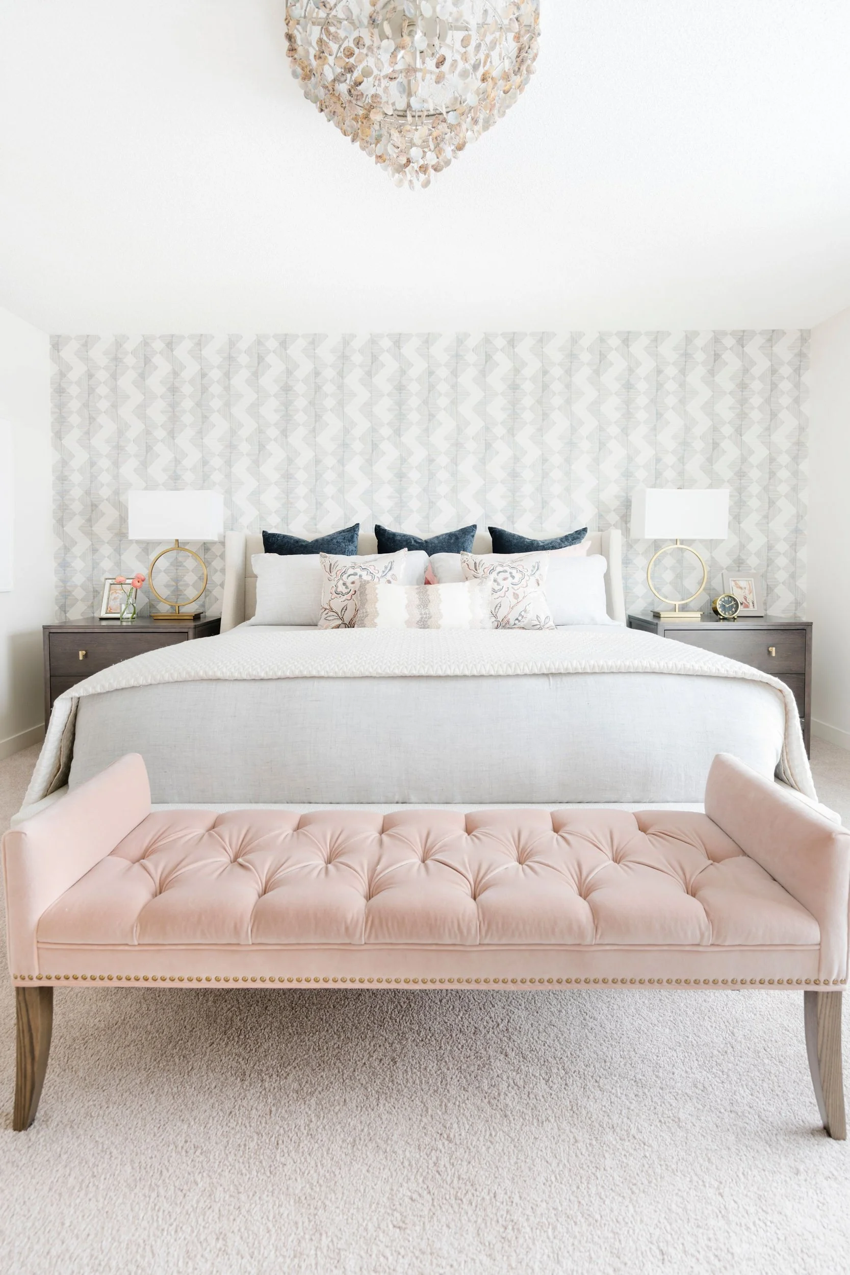 Elegant bedroom with a neutral color palette, featuring a large bed with plush pillows, a tufted pink bench at the foot, and a chic chandelier overhead, set against a patterned wallpaper.