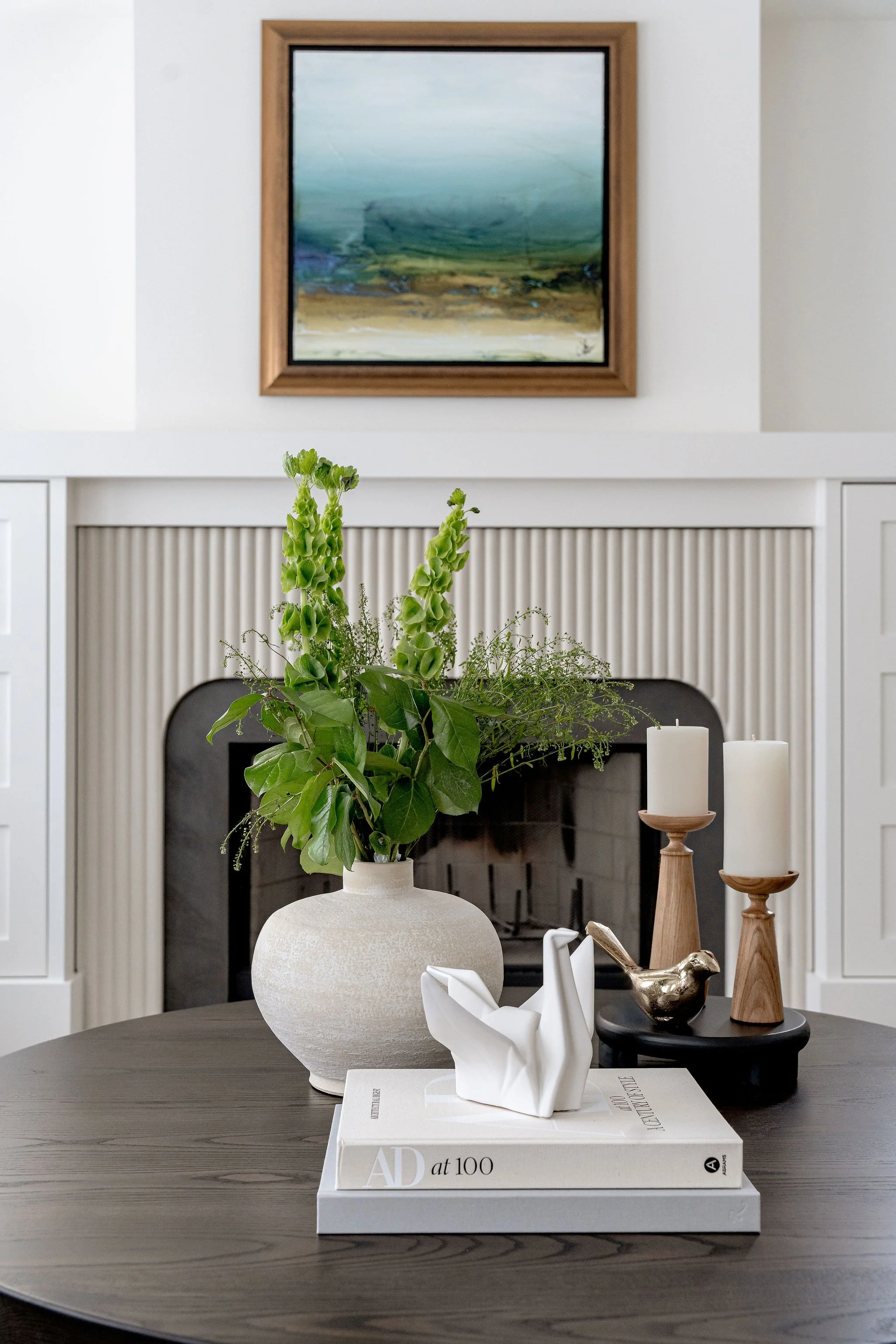Decorative objects on a dining table, including a white ceramic vase with greenery, a white sculpture of a swan on a large book, two wooden candle holders with white candles, and a brass bird figurine. In the background, there is a fireplace with a p