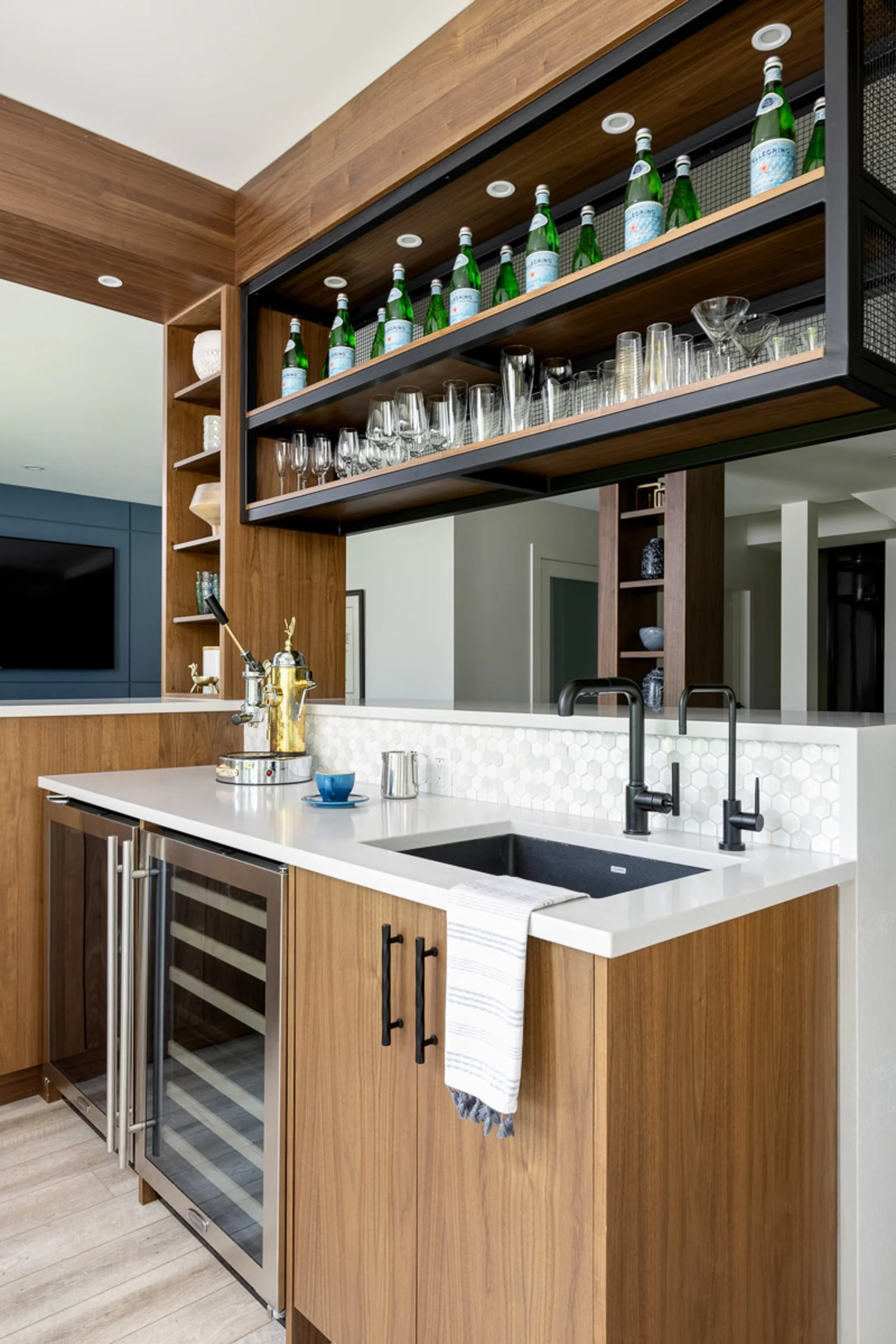 Modern home bar with wood cabinets, glass shelving, and a sink. Features a beverage fridge, espresso machine, glassware, and bottled water. Elegant, sophisticated interior design.