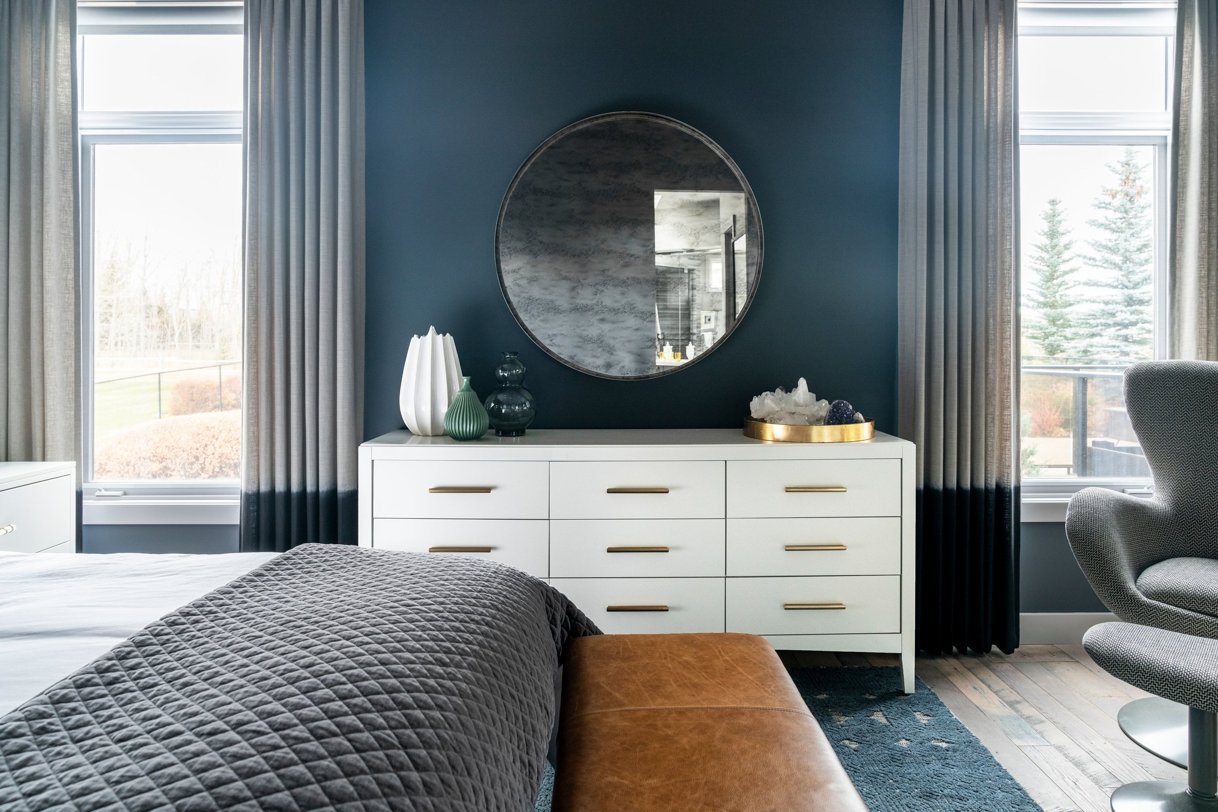 Modern bedroom interior with a white dresser and round mirror. Dark blue walls, large windows with gray curtains, and decorative vases on the dresser. Gray armchair and brown upholstered bench in the foreground.