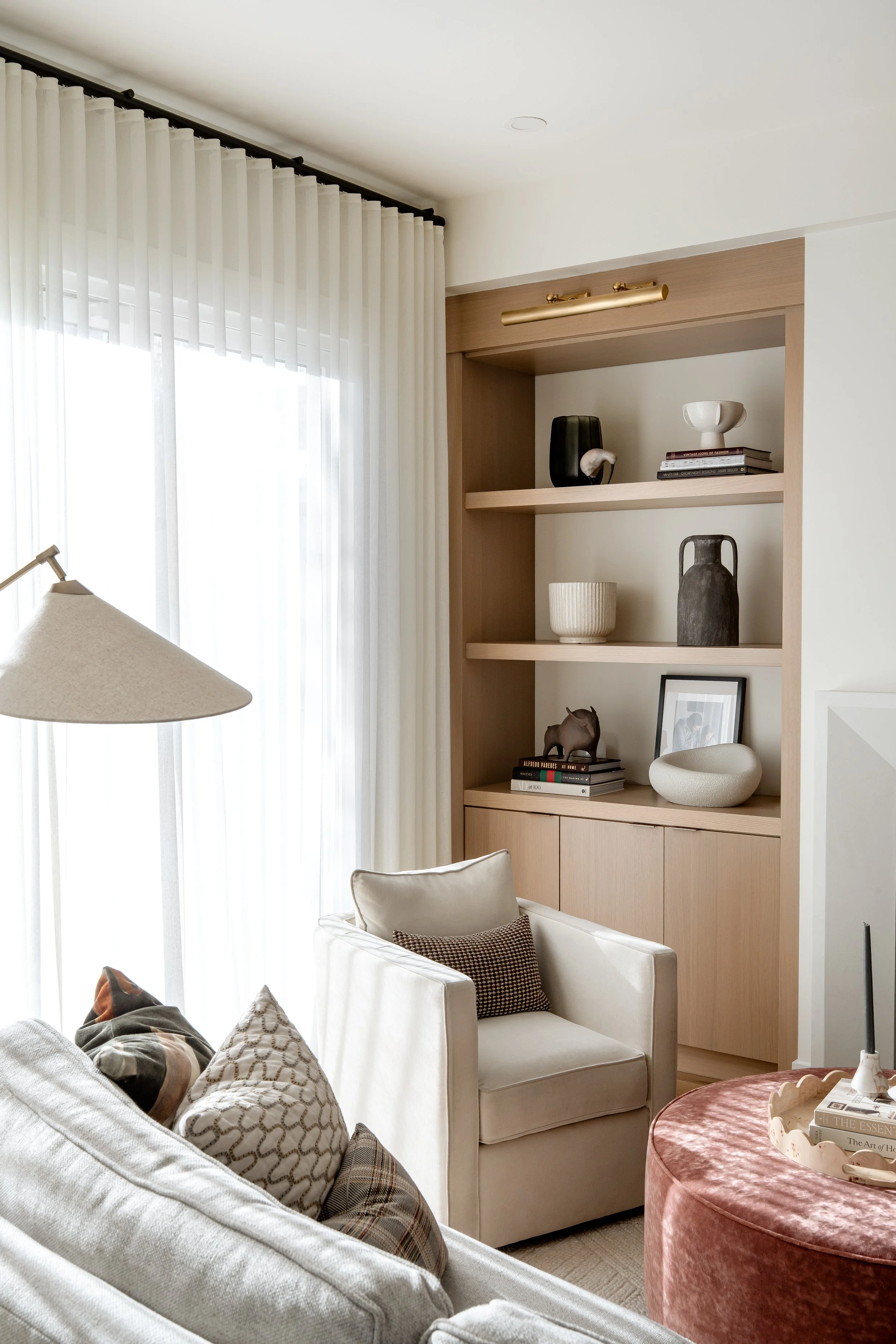 Living room with white sofa, beige armchair, pink round ottoman, bookshelf with decorative items, and large window with sheer curtains.