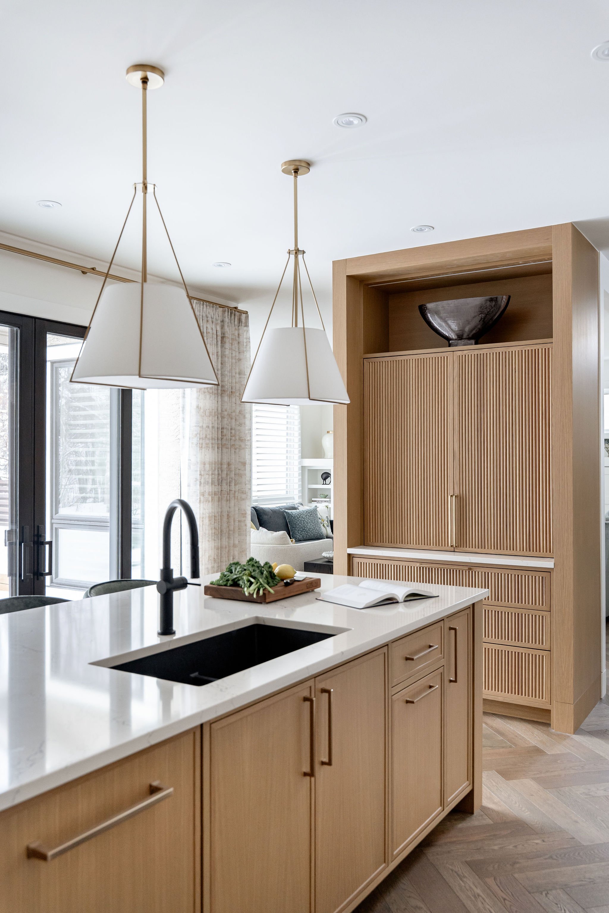 Modern kitchen with light wood cabinets, white island countertop, black sink, and pendant lights, adjacent to living area with window and curtains.