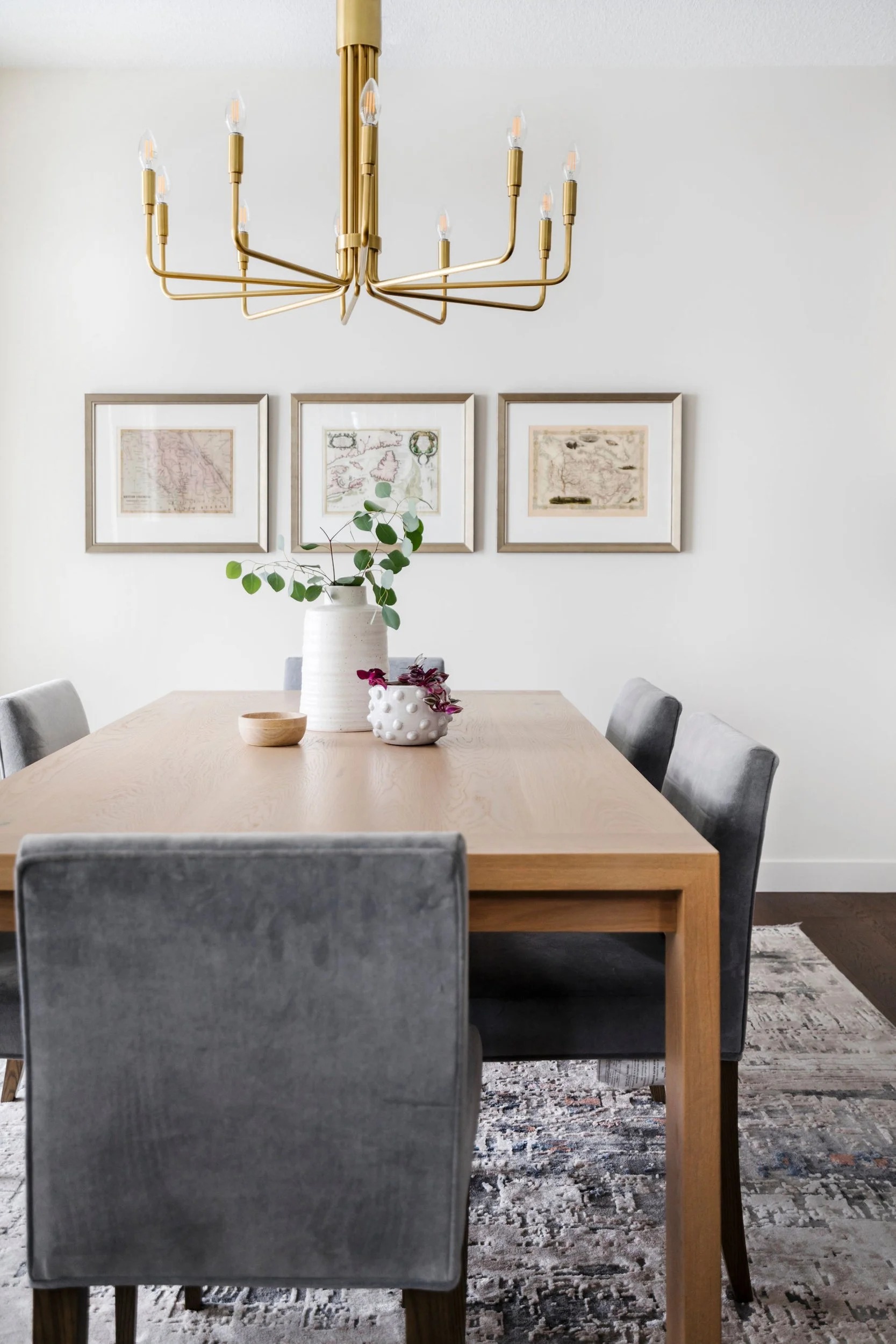 Modern dining room with wooden table, gray chairs, gold chandelier, wall art, and decorative vases with plants.