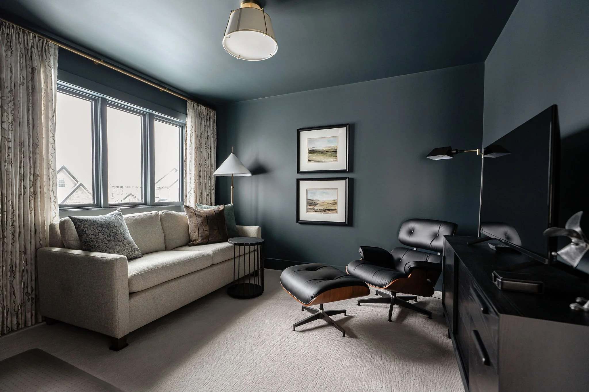 A living room with blue walls, large windows with curtains, a cream-colored sofa with pillows, a small black round table, framed artwork on the wall, a white floor lamp, and a black swivel chair near a black desk with a TV.
